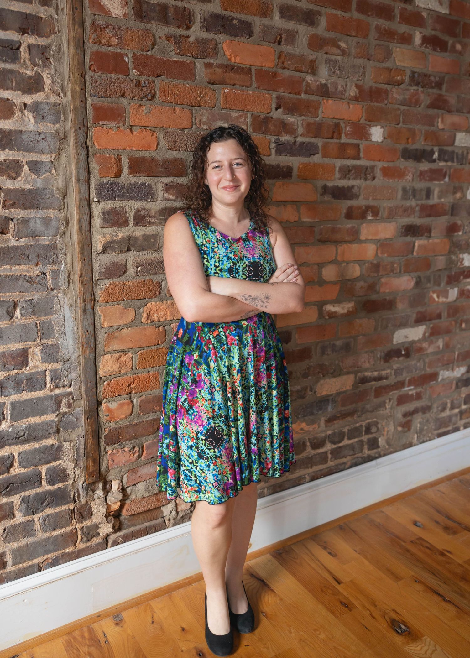 Person in a colorful floral print dress poses with crossed arms against a rustic exposed brick wall on hardwood floors.