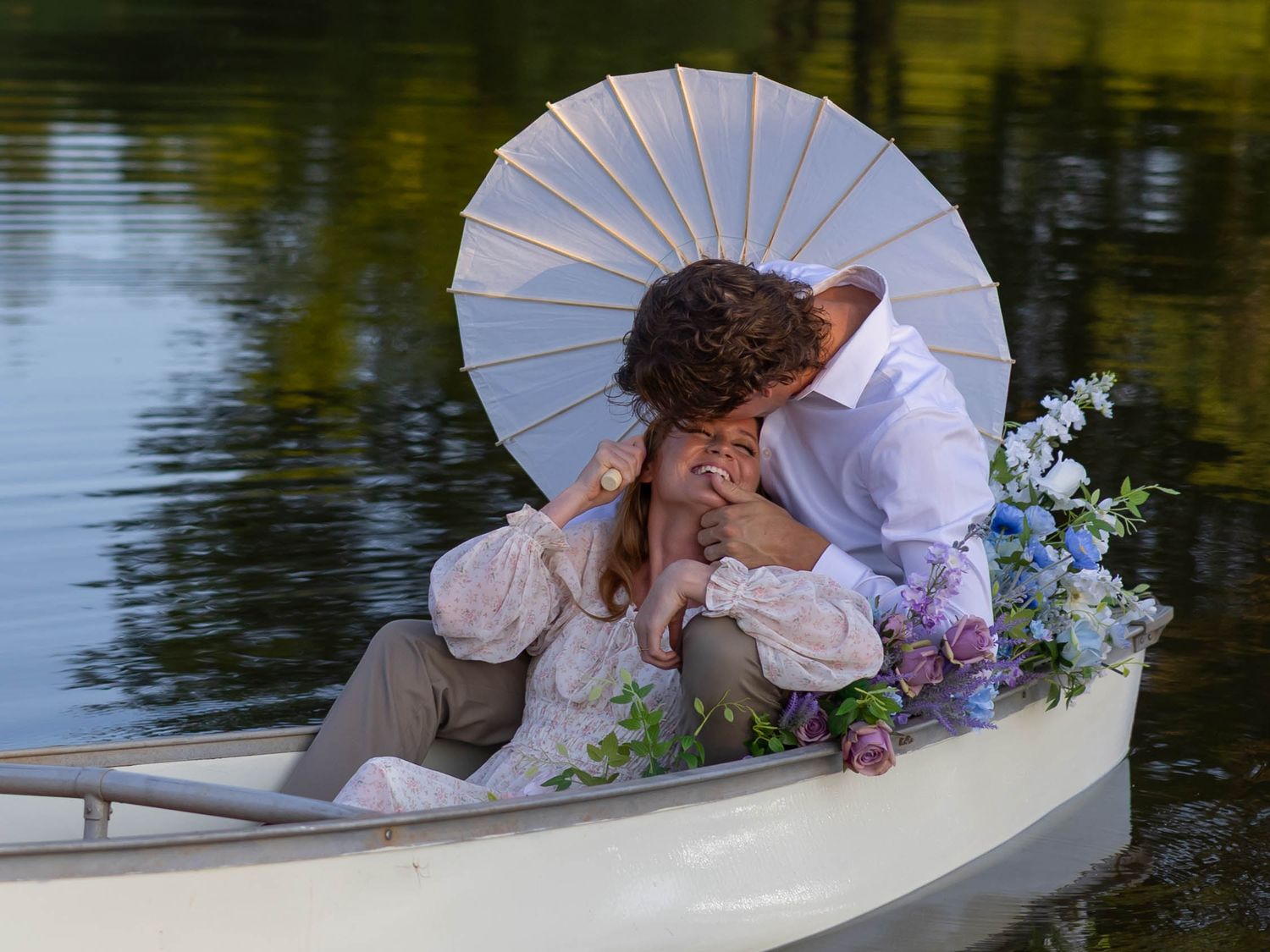 A romantic couple in wedding attire shares tender moments while sitting in a white rowboat on a serene lake.