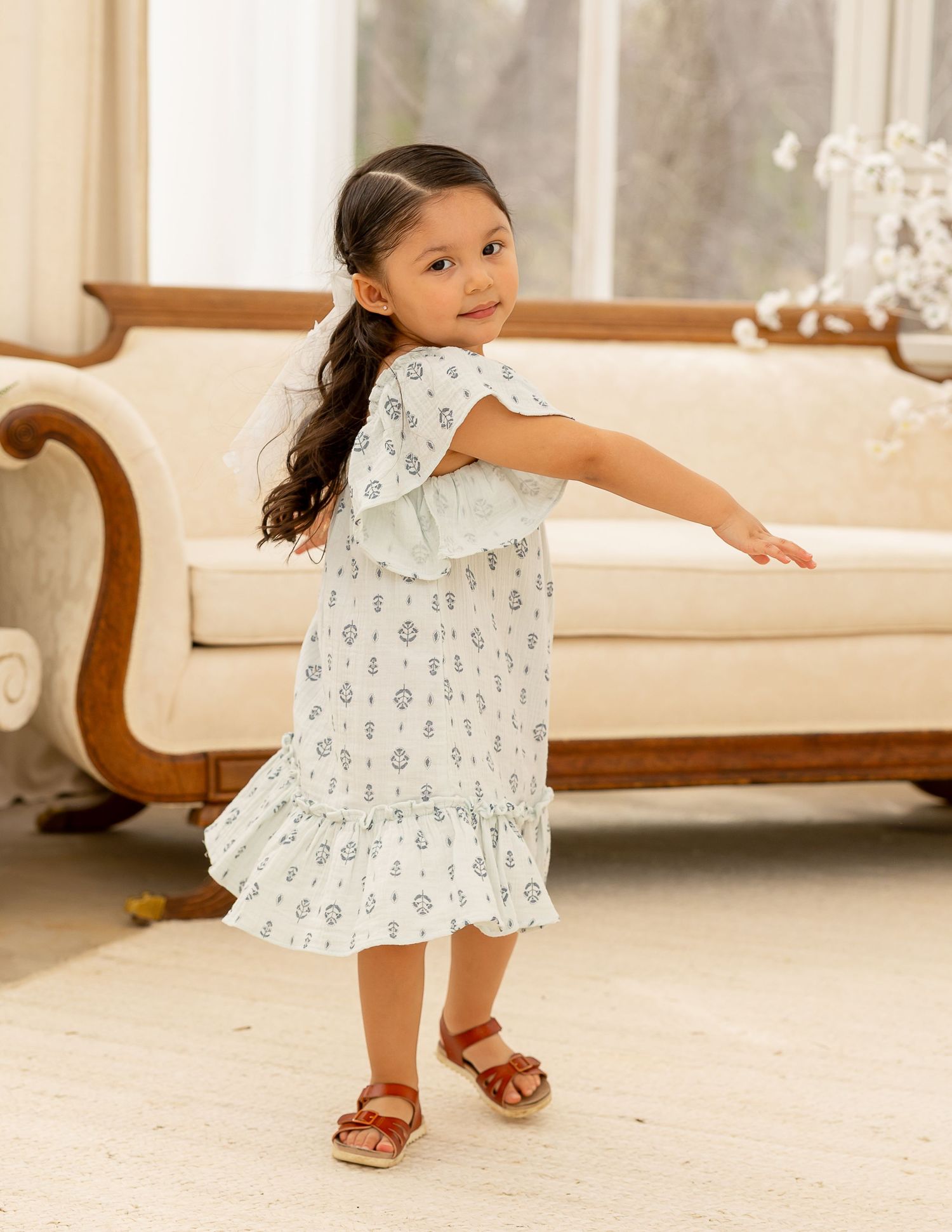 Young girl in white floral dress twirls and dances on cream carpet during indoor photo session.