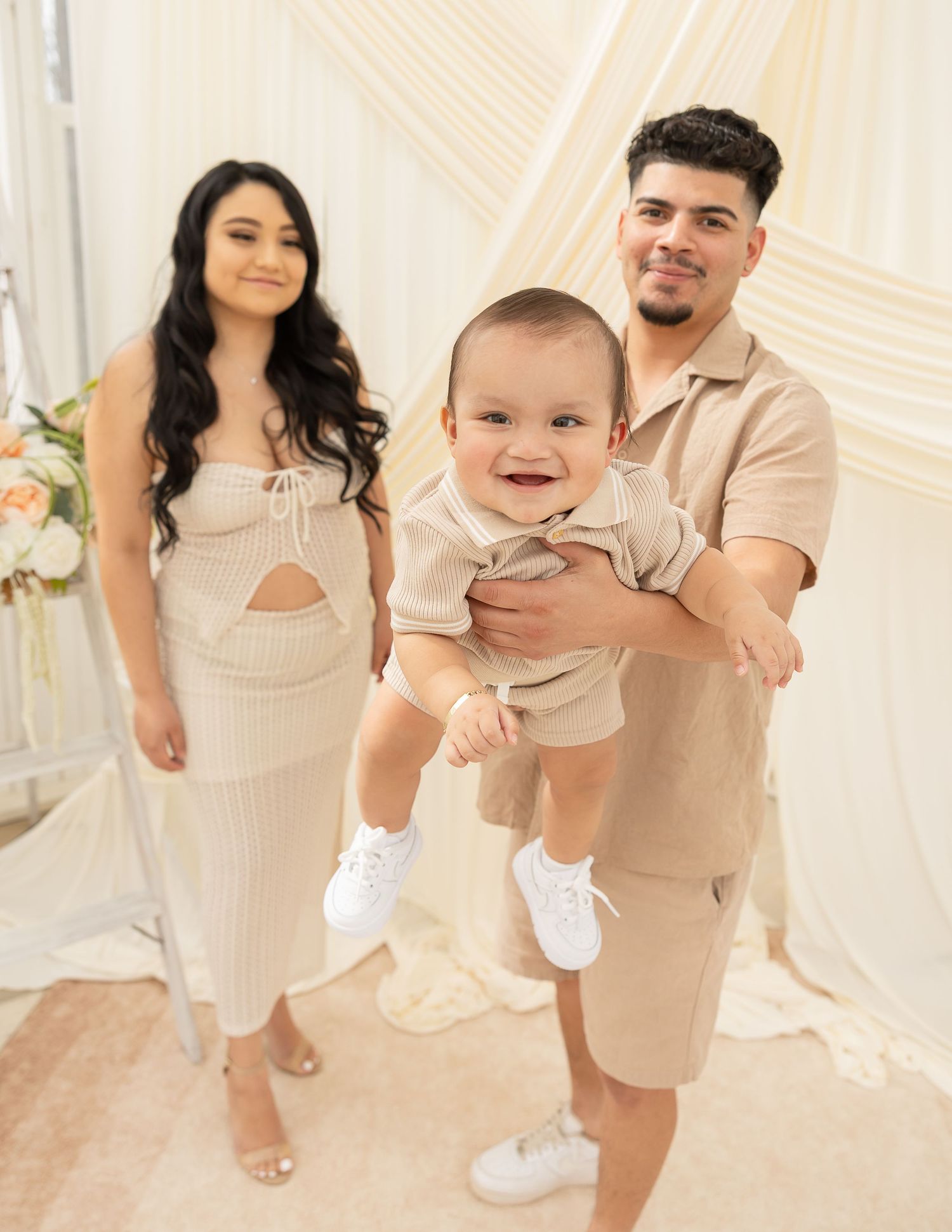 A family wearing coordinated beige and white outfits poses together in a bright studio setting for a series of photos.