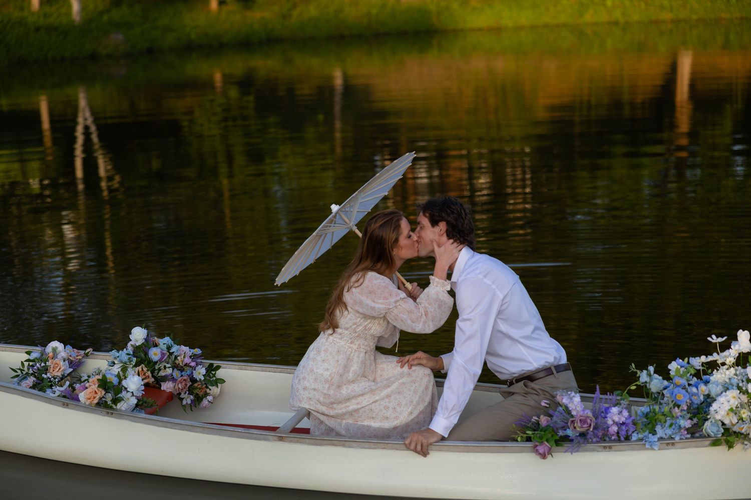 Couple in white wedding attire share a romantic moment on a wooden dock surrounded by colorful floral arrangements.