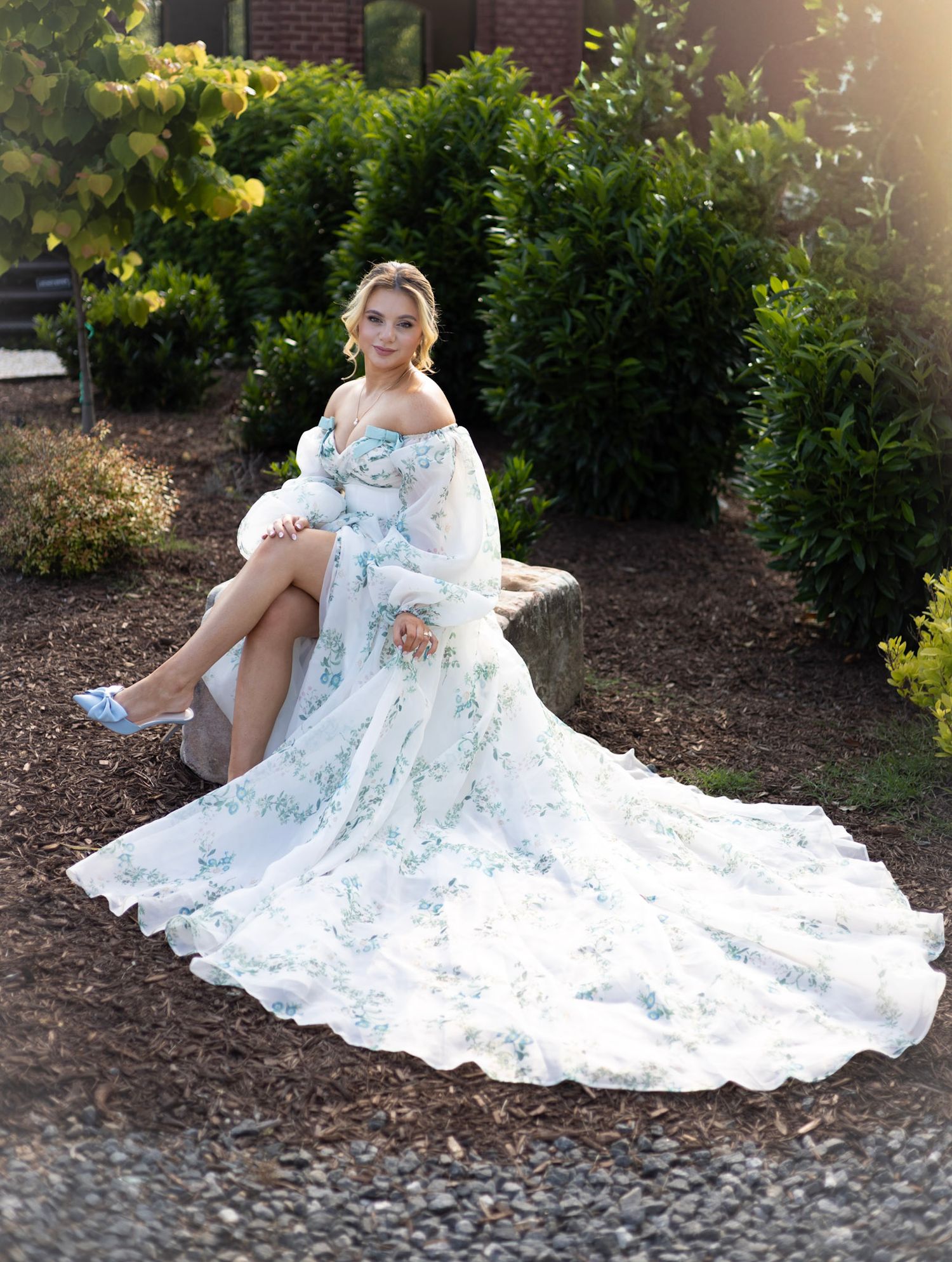 A flowing white floral wedding dress spreads elegantly across mulched garden ground in outdoor portrait session.