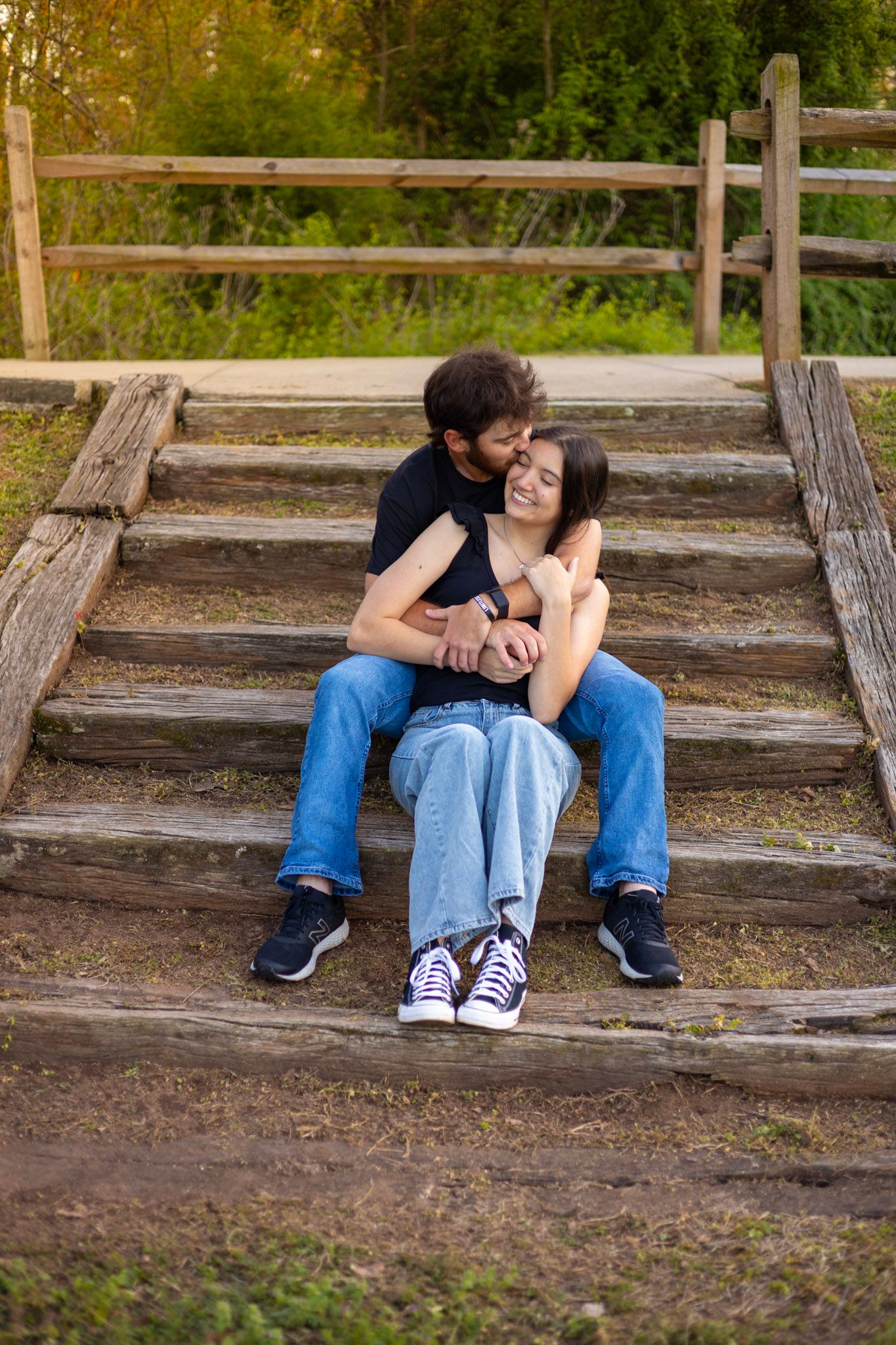 A couple sits close together on rustic wooden steps outdoors, sharing an intimate moment in a natural setting.
