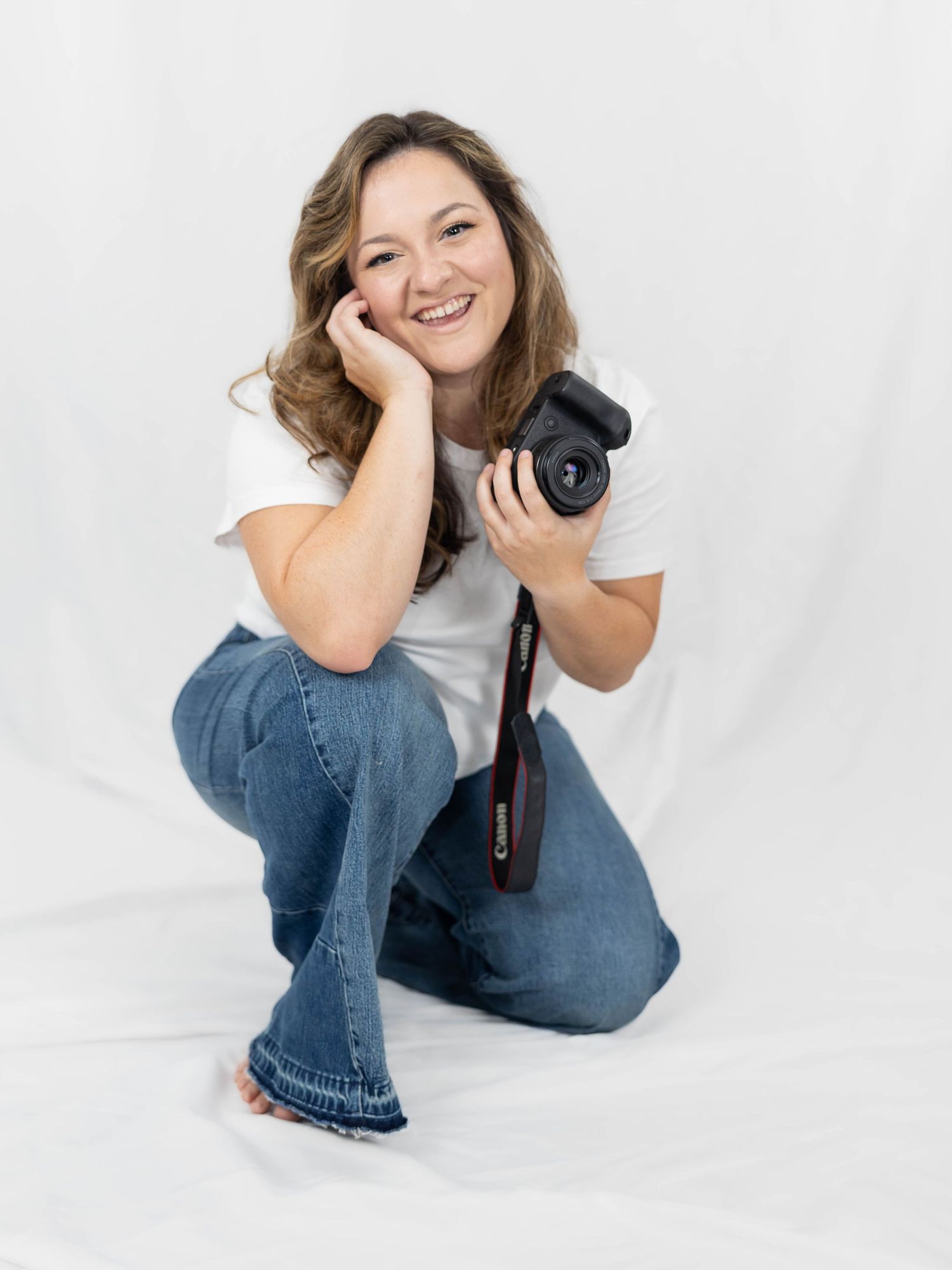 Professional photographer crouching with a camera in hand wearing a white t-shirt and jeans against white backdrop.