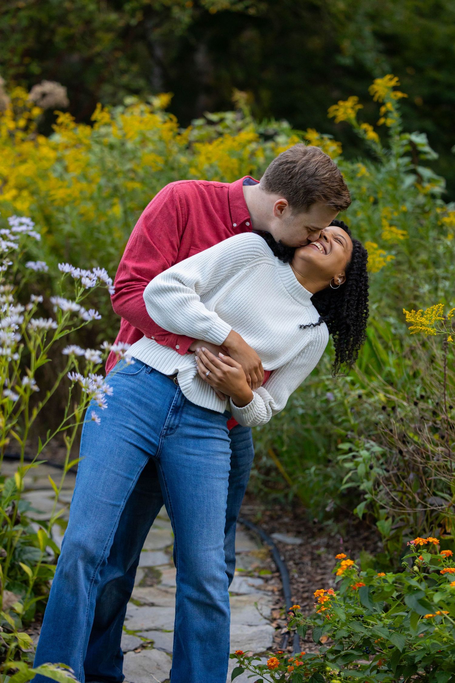 A couple embraces in front of vibrant yellow wildflowers along a garden path on a sunny day.