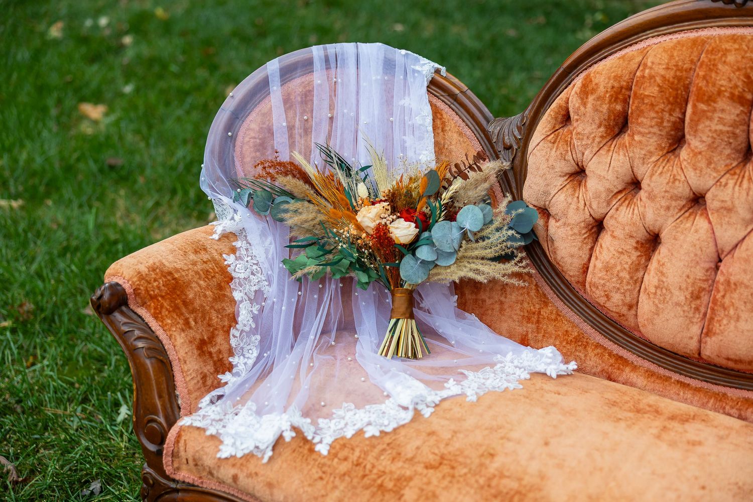 Bridal bouquet with dried flowers and eucalyptus resting on a white lace veil draped over a vintage velvet tufted sofa.