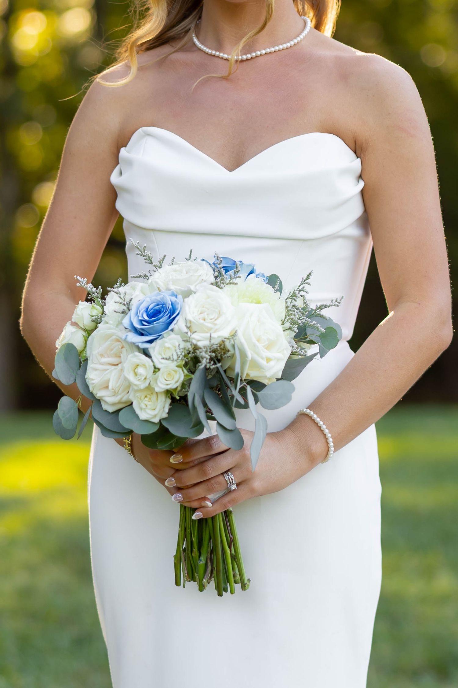 Bride in strapless white gown holding bouquet of white and blue roses with eucalyptus, wearing pearl necklace and bracelet.