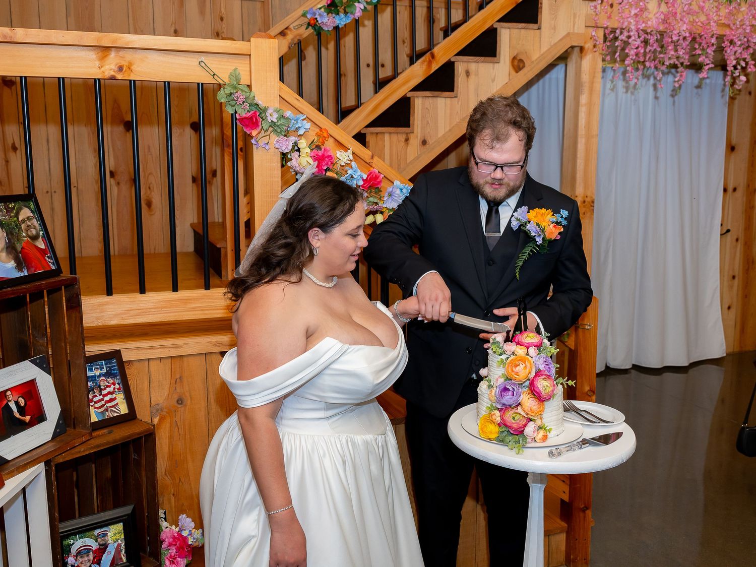 Bride and groom cut their colorful floral wedding cake inside a rustic wood-paneled venue.
