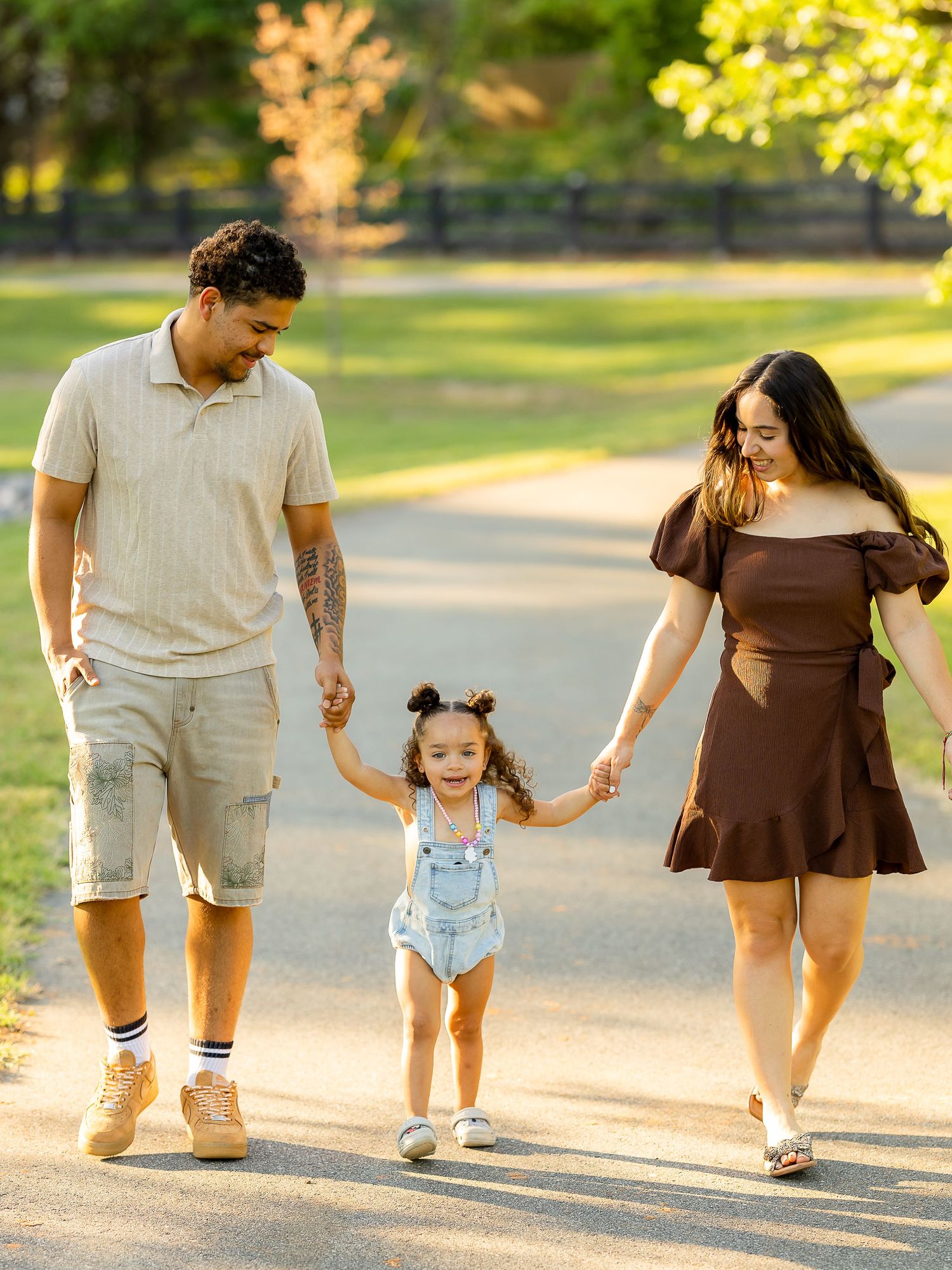 A happy family of three walks hand-in-hand in a sunny park, parents swinging their toddler girl in denim overalls.