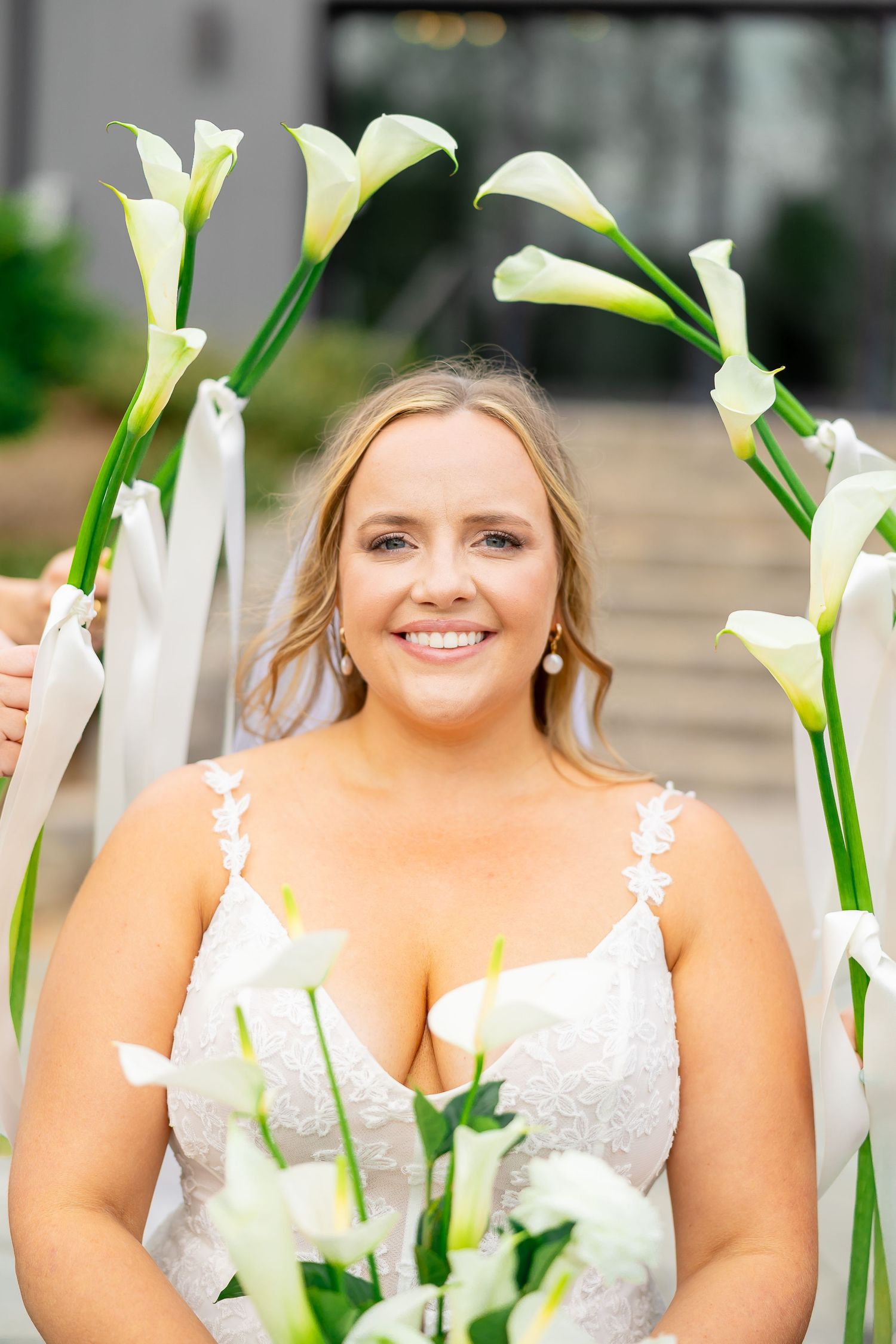 Smiling bride holding white calla lilies bouquet framed by elegant floral arch in lace wedding gown.