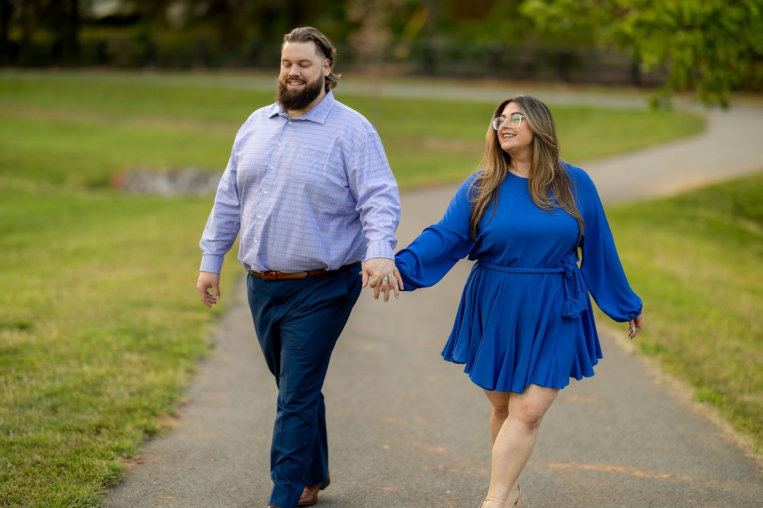 Couple holding hands walking on a park path, man in blue shirt and woman in royal blue dress, outdoor engagement photo.