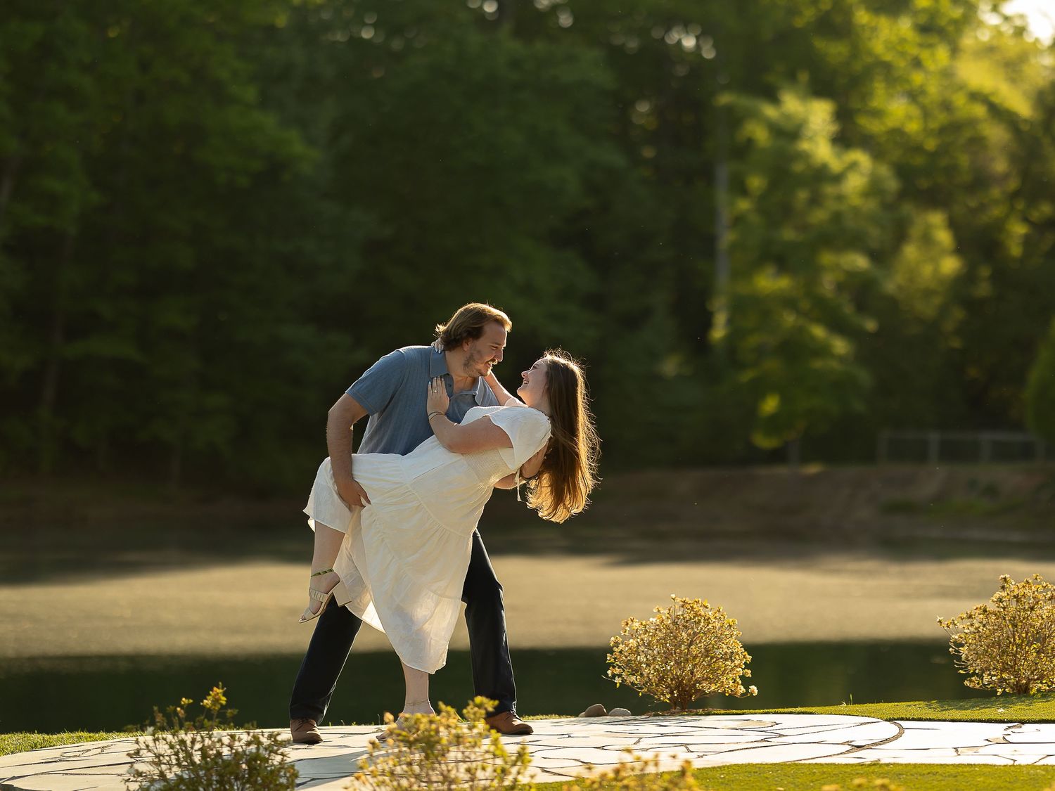 Couple sharing romantic dip kiss on stone patio surrounded by lush green trees and pond at golden hour.