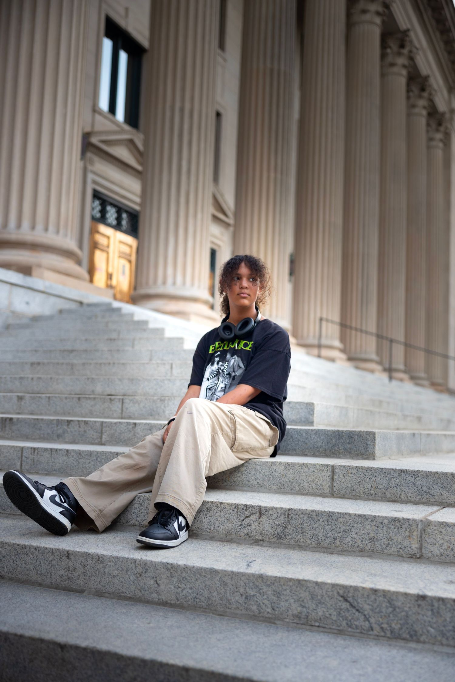 A person sits casually on gray stone steps wearing khaki pants, black t-shirt, and sneakers near classical columns.