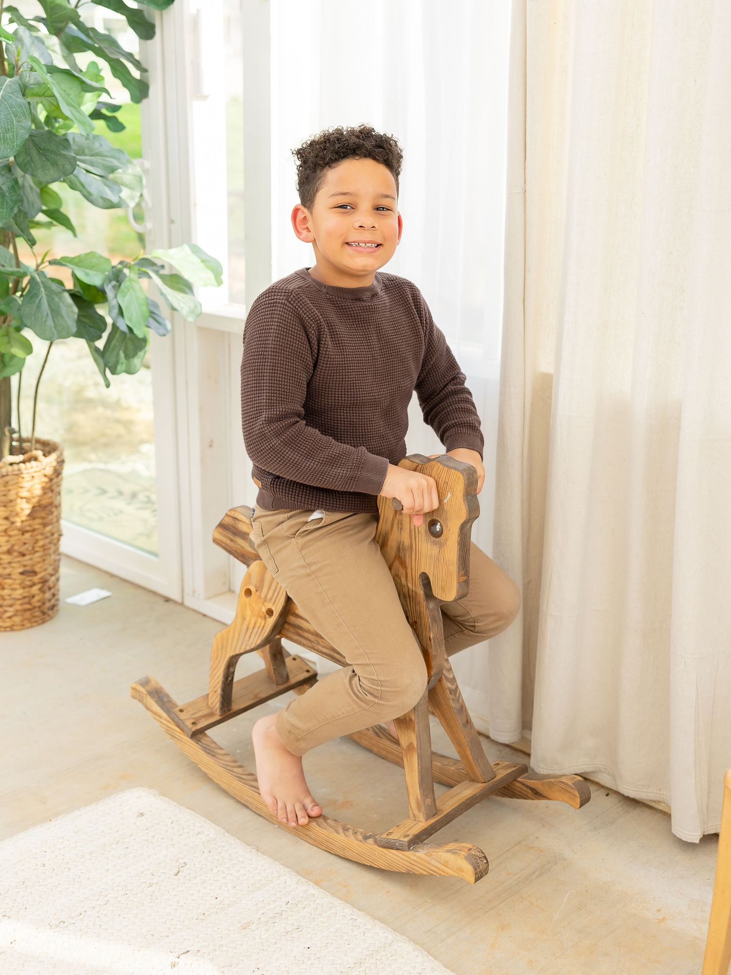 Smiling boy in brown sweater and khaki pants sitting on a wooden rocking horse near a bright window.