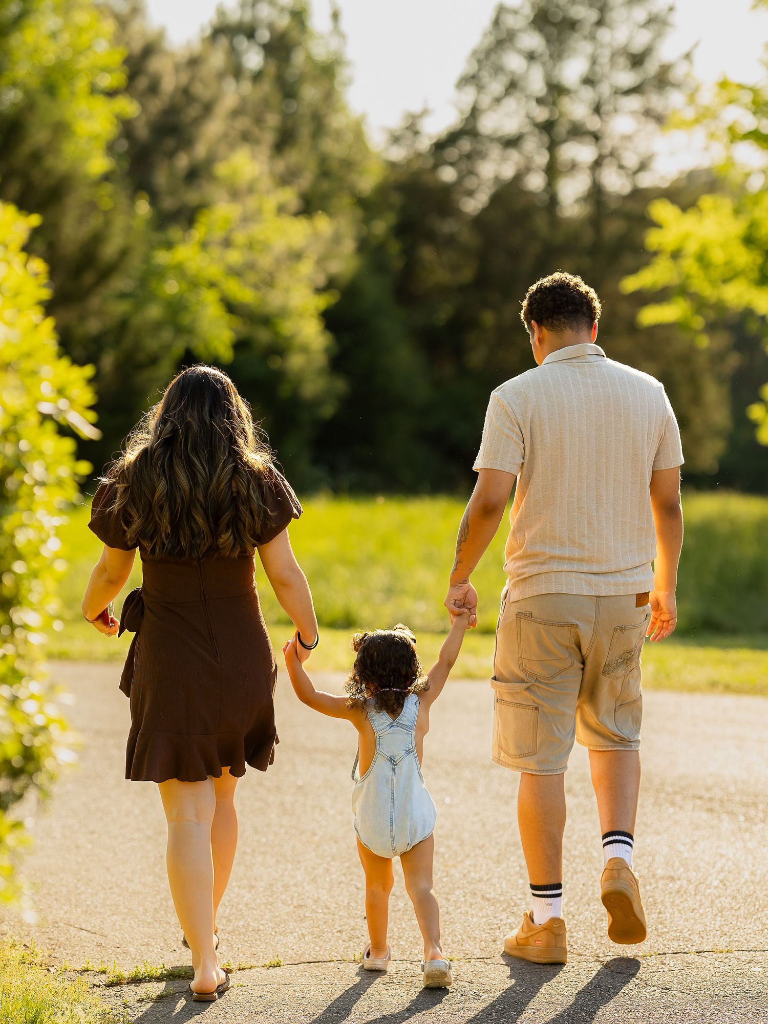 A family of three walks hand-in-hand down a sunlit park path, parents in brown dress and khaki shorts with toddler between them.