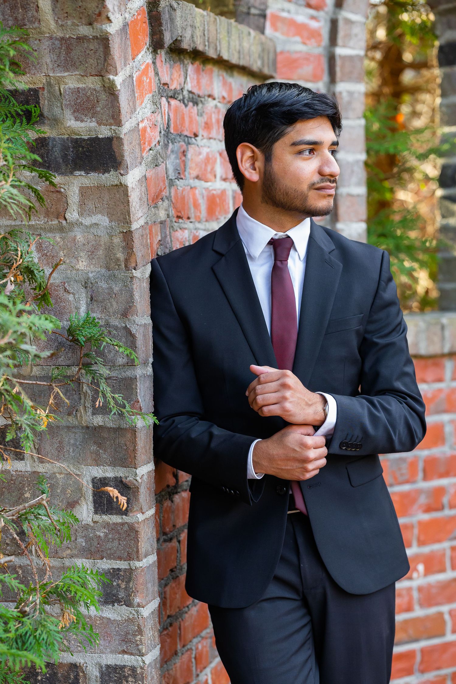Young man in black suit and burgundy tie leaning against brick wall outdoors.