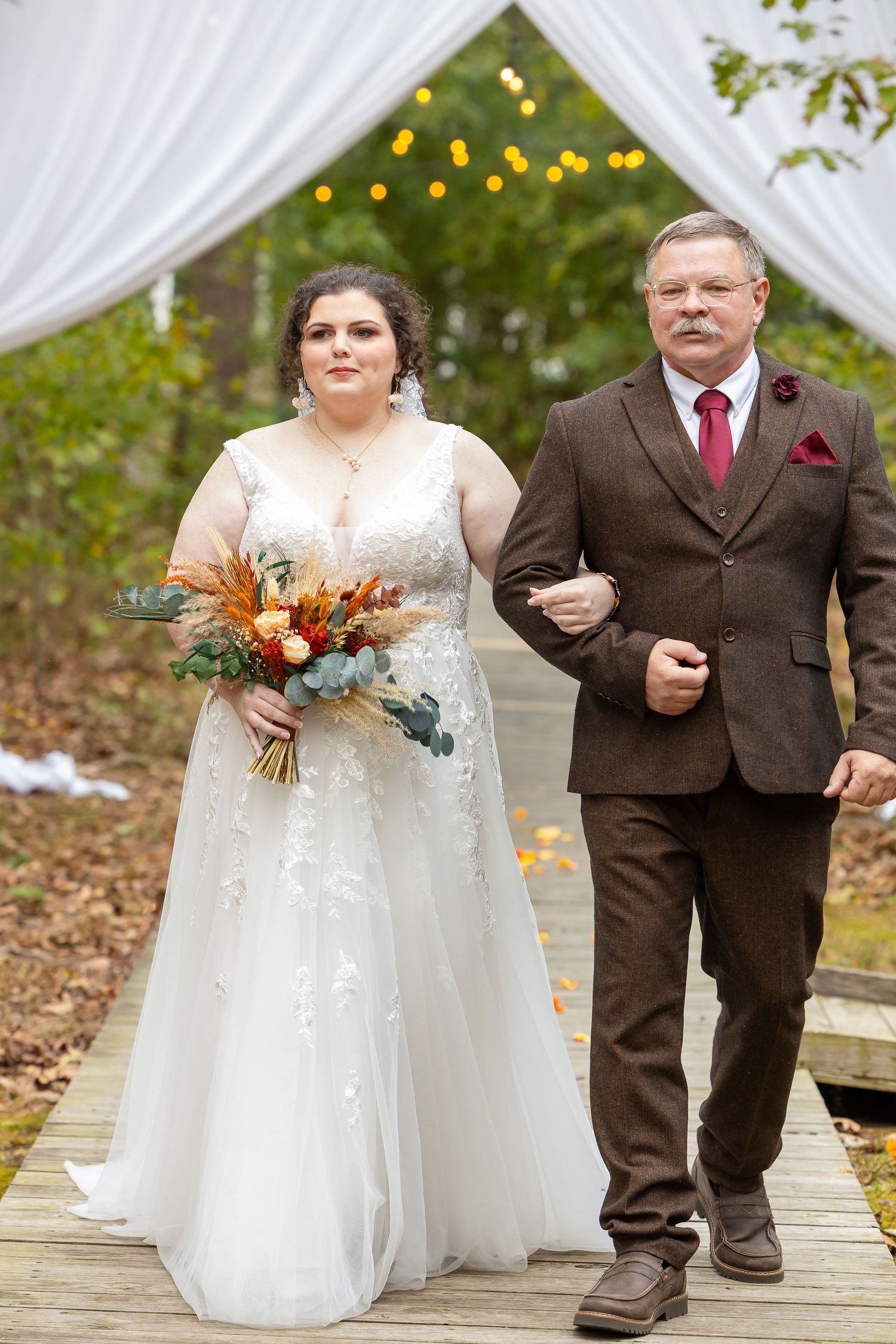 Bride in white lace gown holding autumn bouquet walks down outdoor aisle with escort in brown suit.
