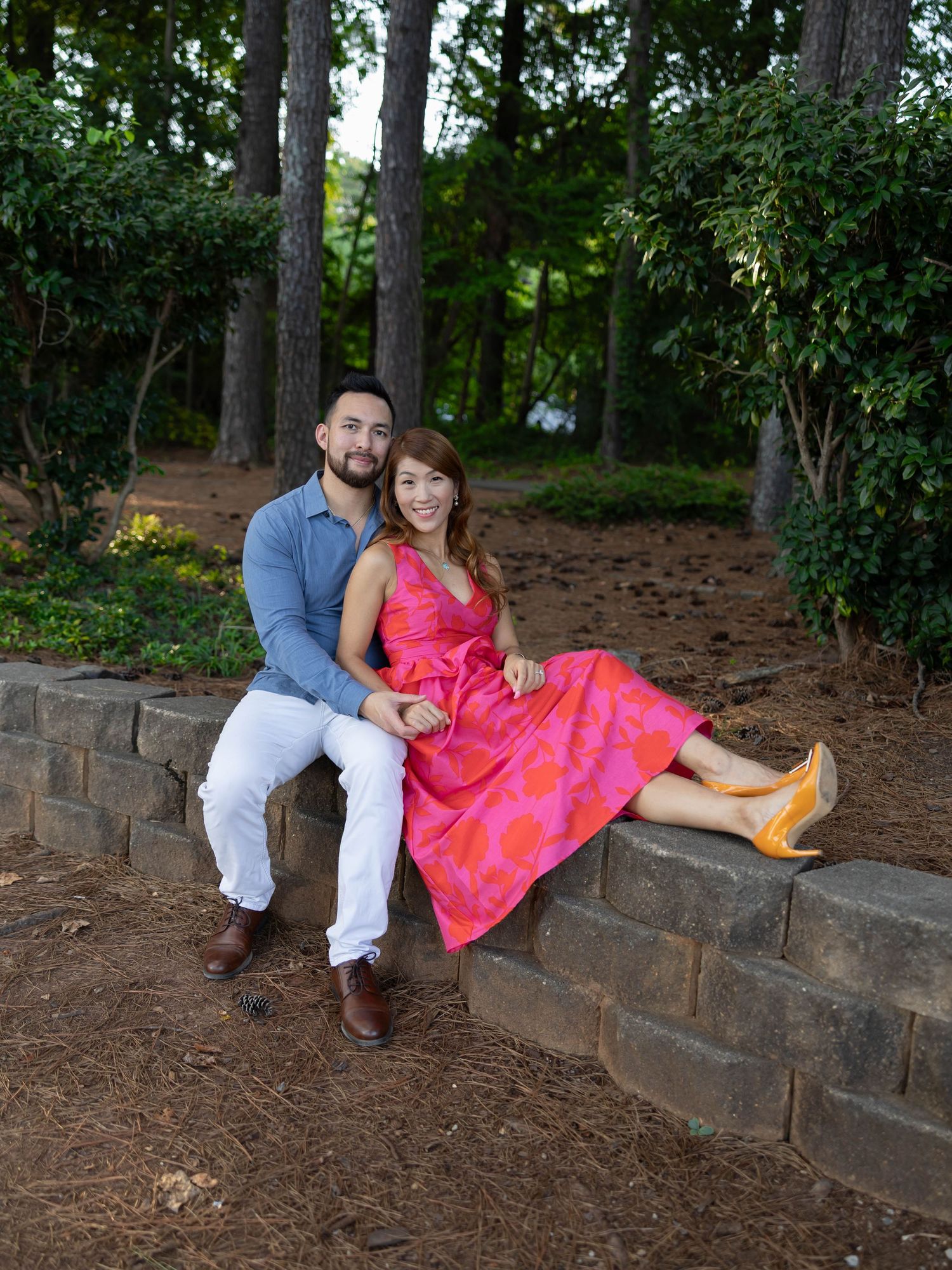A couple in romantic attire sits together on stone retaining wall steps in a wooded outdoor setting.