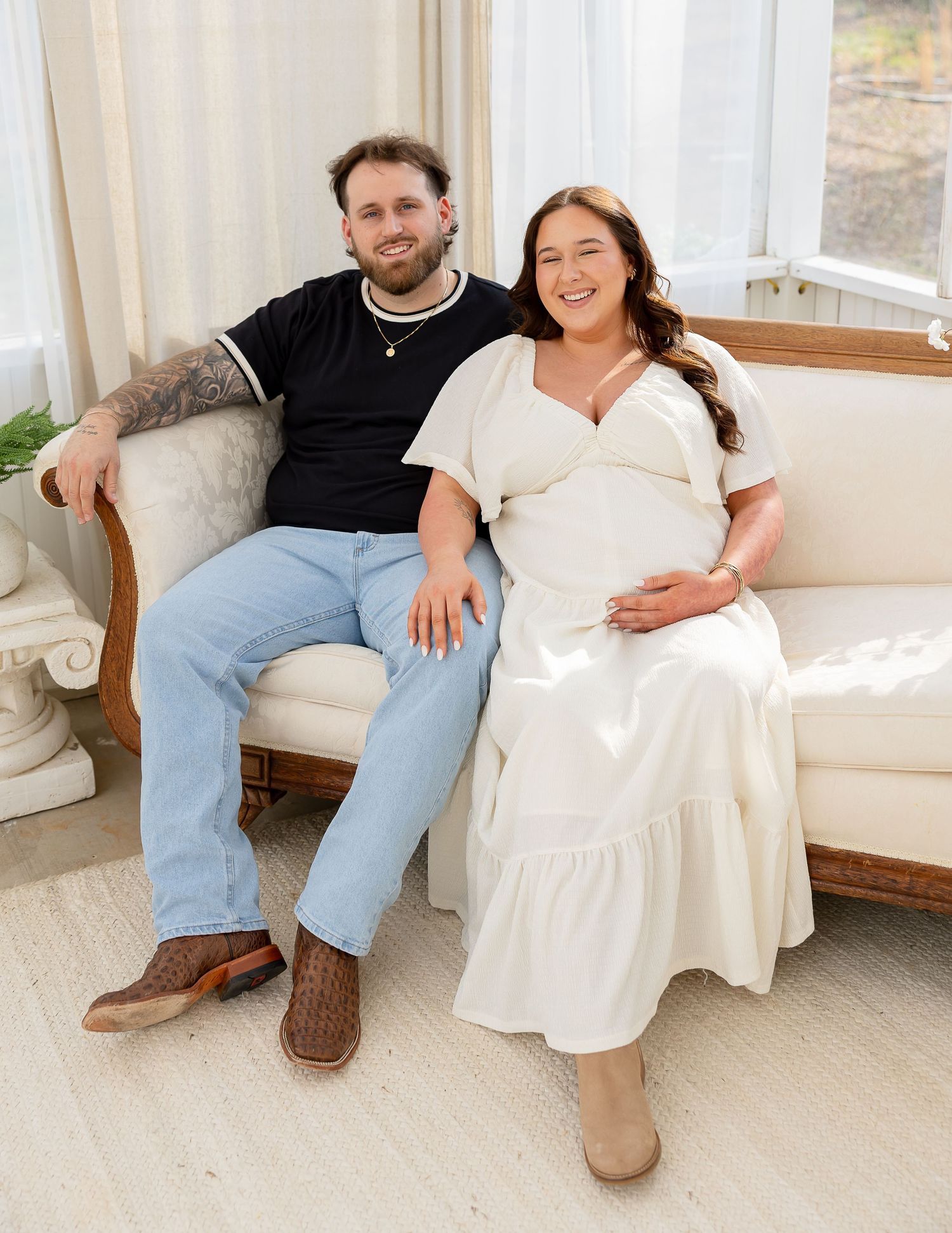 A couple sits casually together on a white bench in a bright room wearing jeans and white clothing.