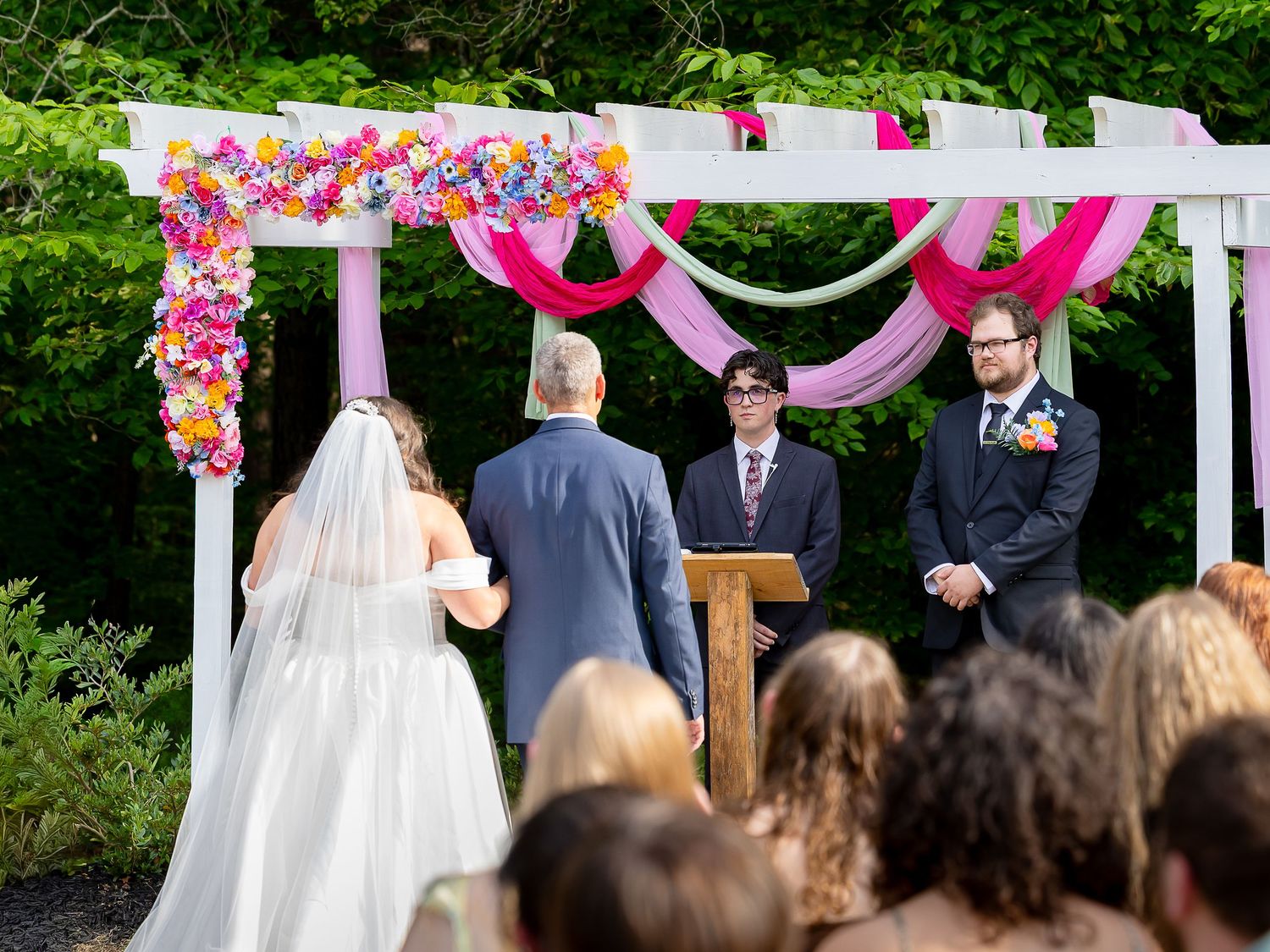 Outdoor wedding ceremony under floral arch with pink and colorful flowers as bride walks toward groom and officiant.