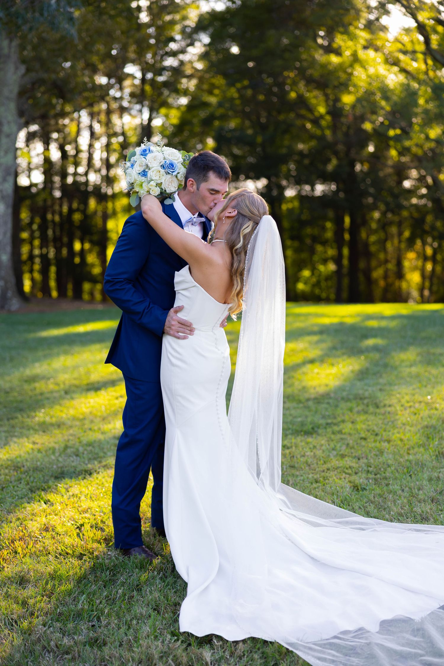 Bride and groom share a romantic kiss outdoors on green grass with trees in background on their wedding day.