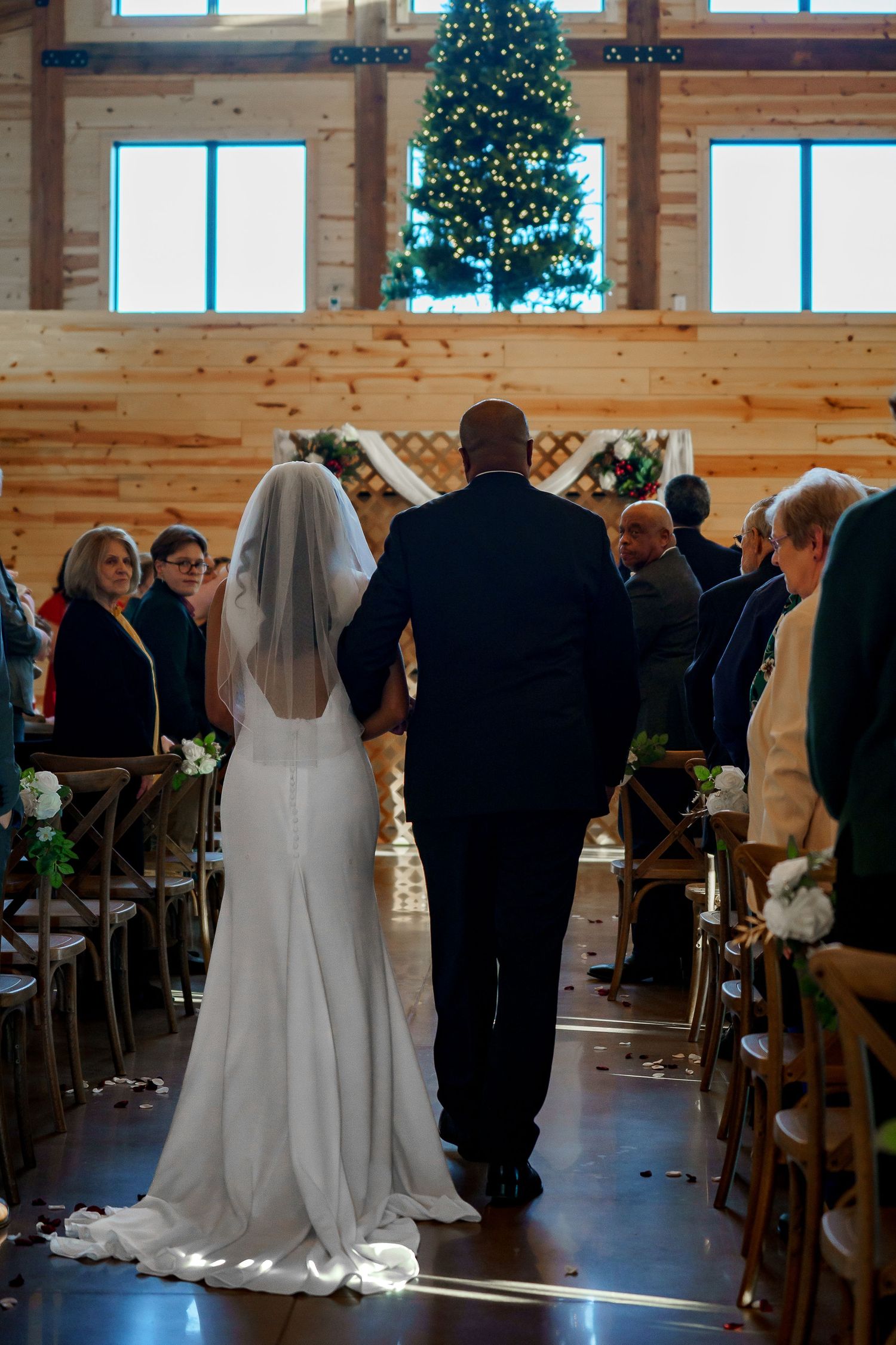 Bride in white gown and veil walks down aisle with escort in rustic barn wedding venue with Christmas tree.