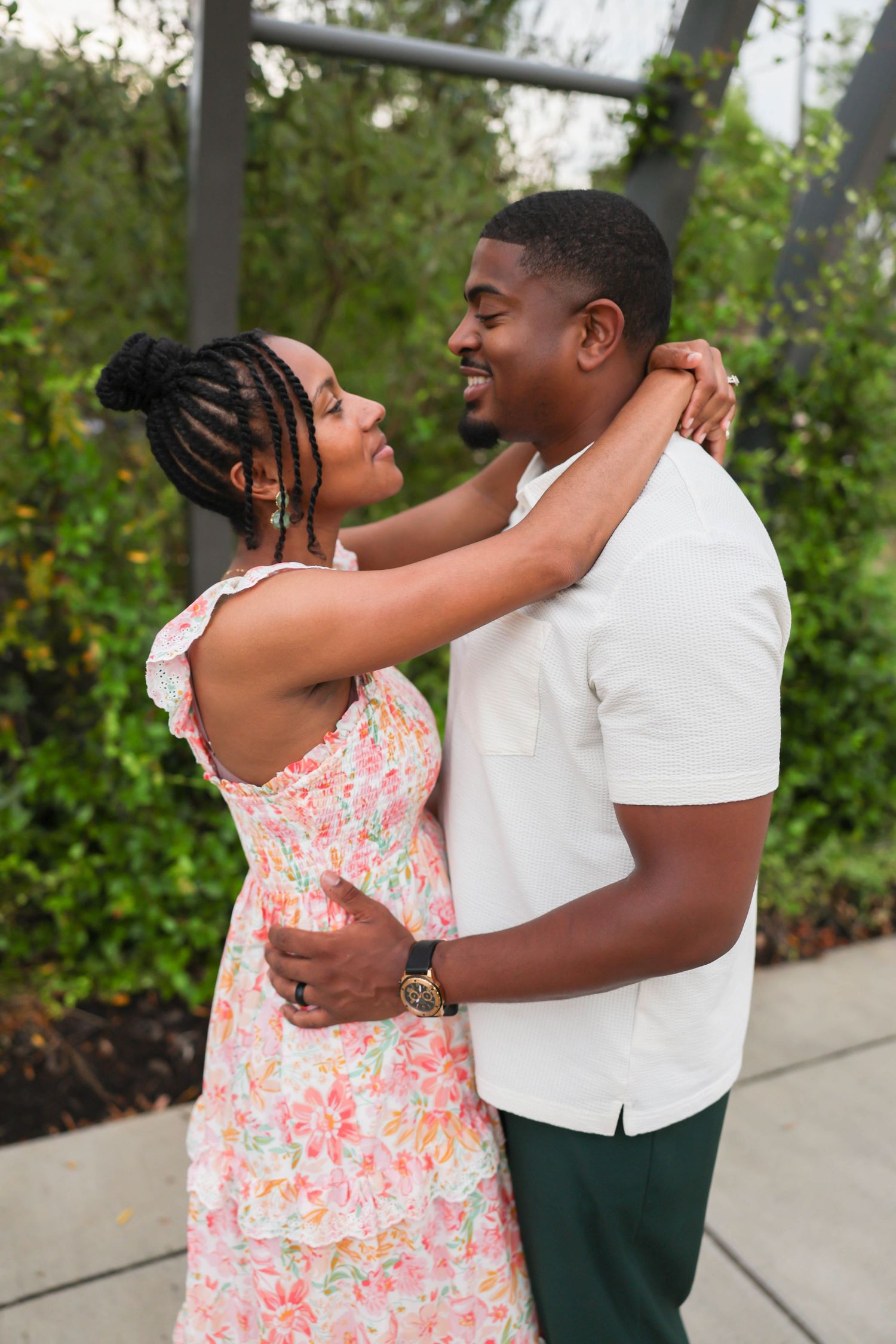 A happy couple embraces outdoors, with her wearing a floral sundress and him in a white polo and green pants.