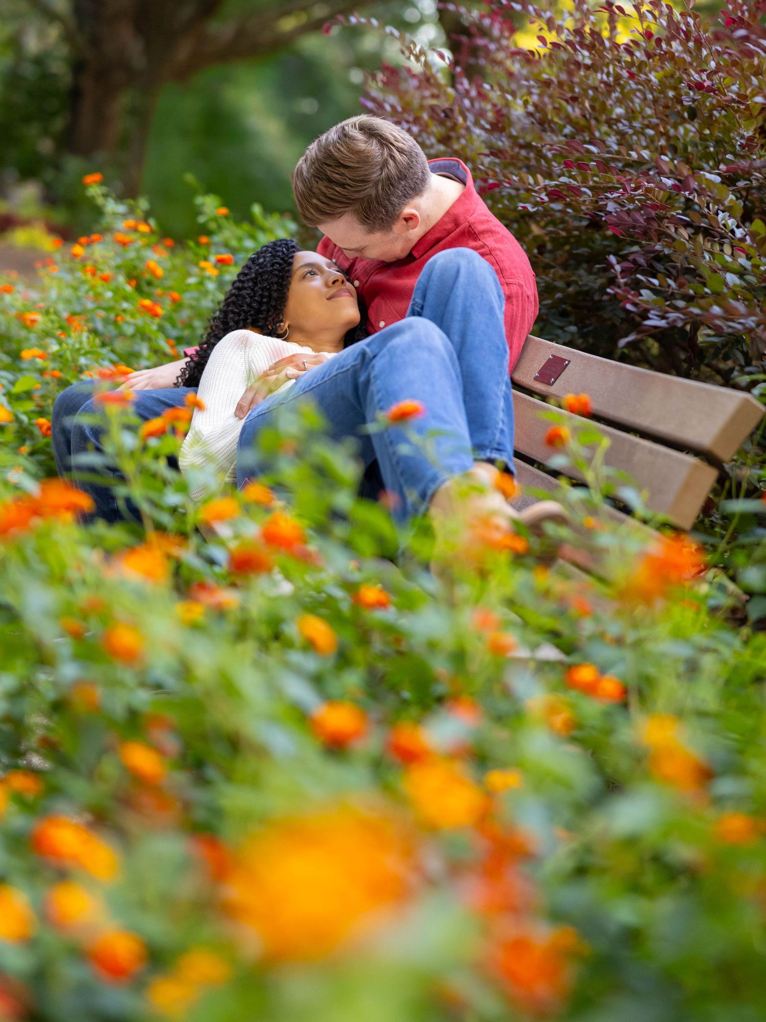 Couple shares tender moment together on park bench surrounded by vibrant orange and green garden flowers.