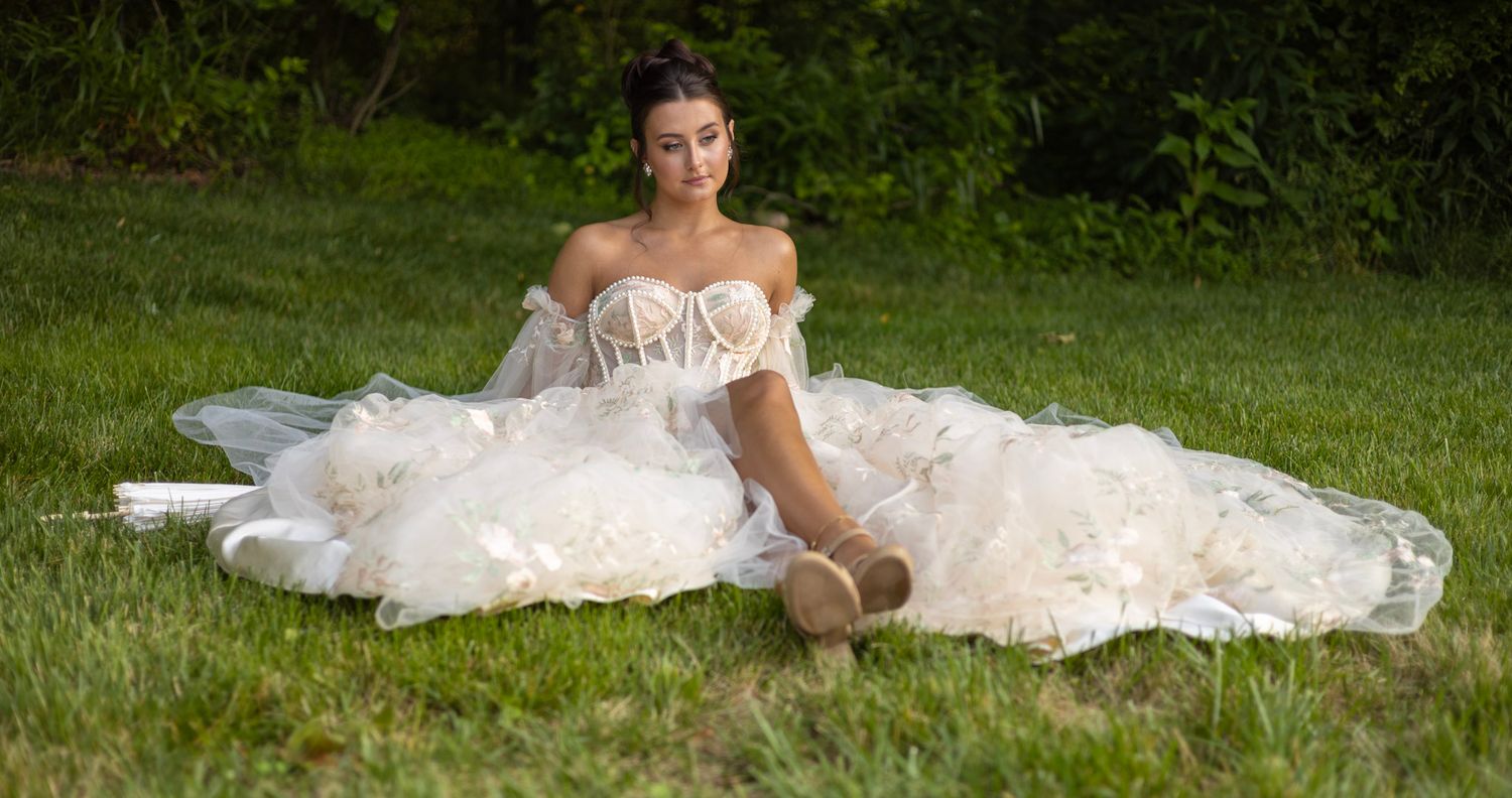 A bride in a flowing white wedding gown relaxes on lush green grass during an outdoor photo session.