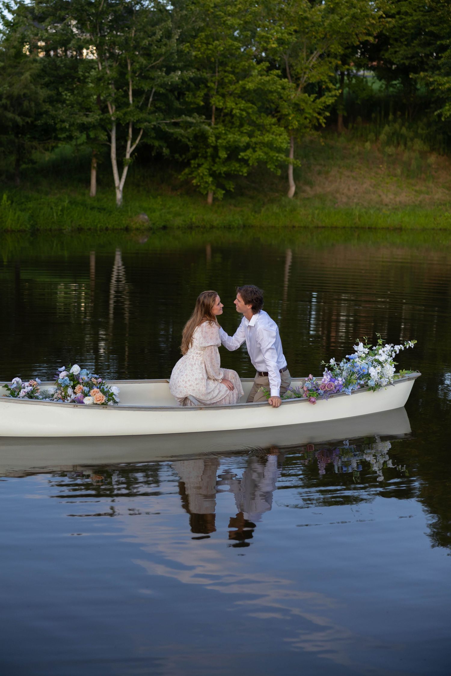 A romantic couple shares a tender moment in a white canoe decorated with flowers on a serene lake at sunset.