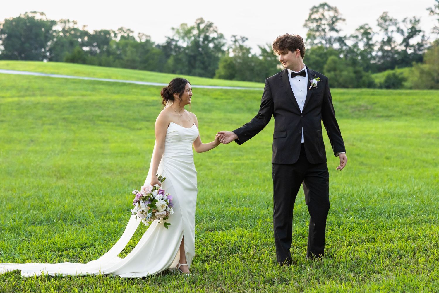 A bride and groom hold hands while walking together on lush green grass during their outdoor wedding.