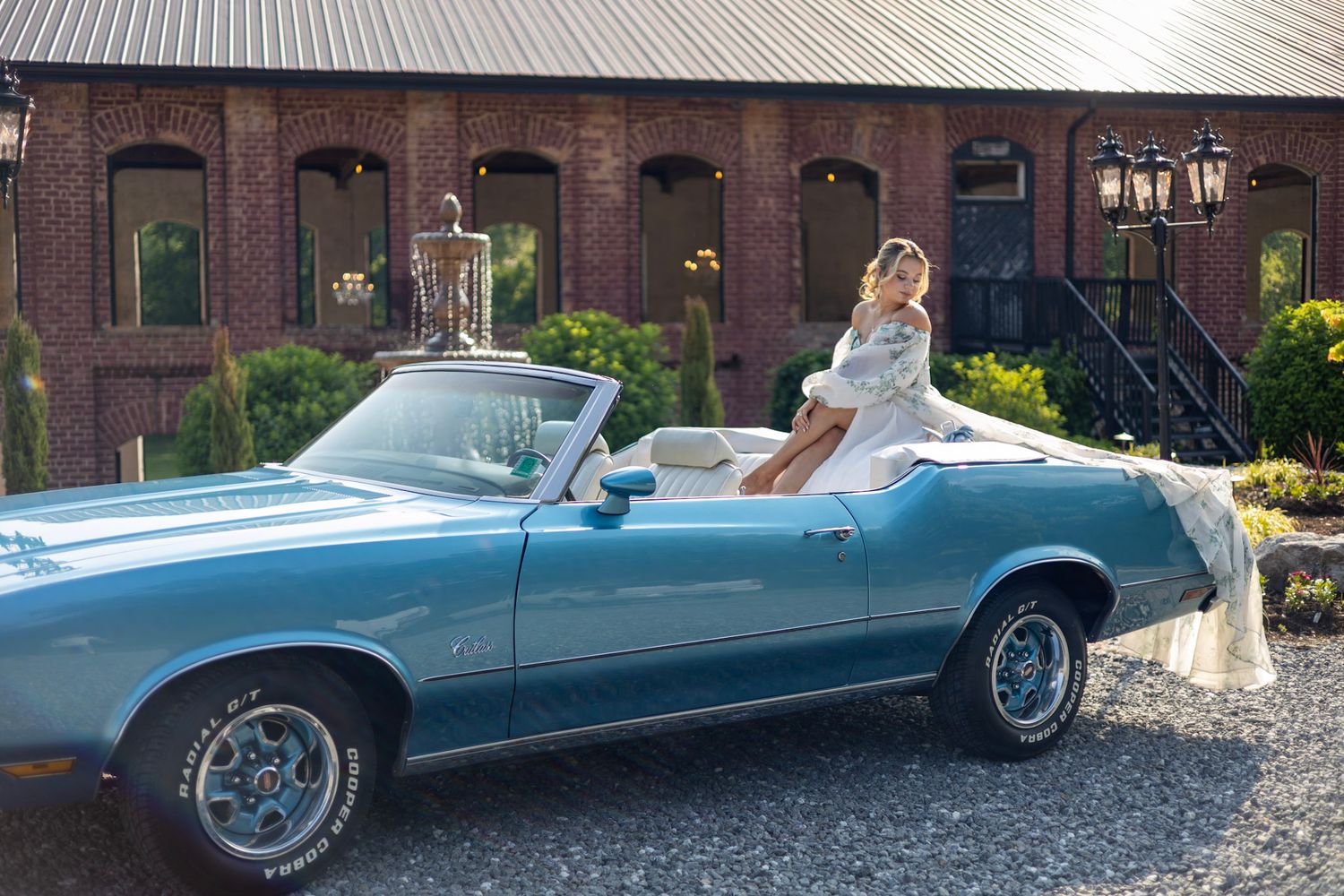 A classic blue convertible car parked in front of a brick building with a person seated in the driver's seat.
