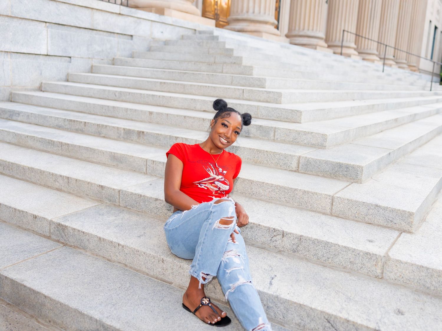 Person in red t-shirt and ripped jeans sitting on white marble steps with architectural columns in background.