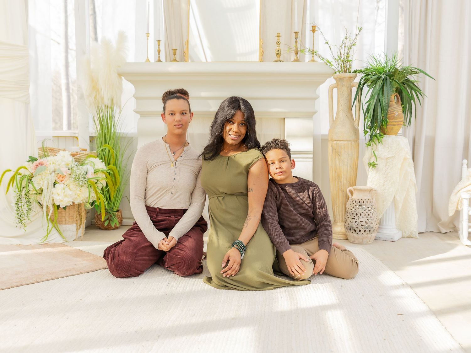A woman and two children pose together on the floor in front of a white fireplace in a bright, elegant room.