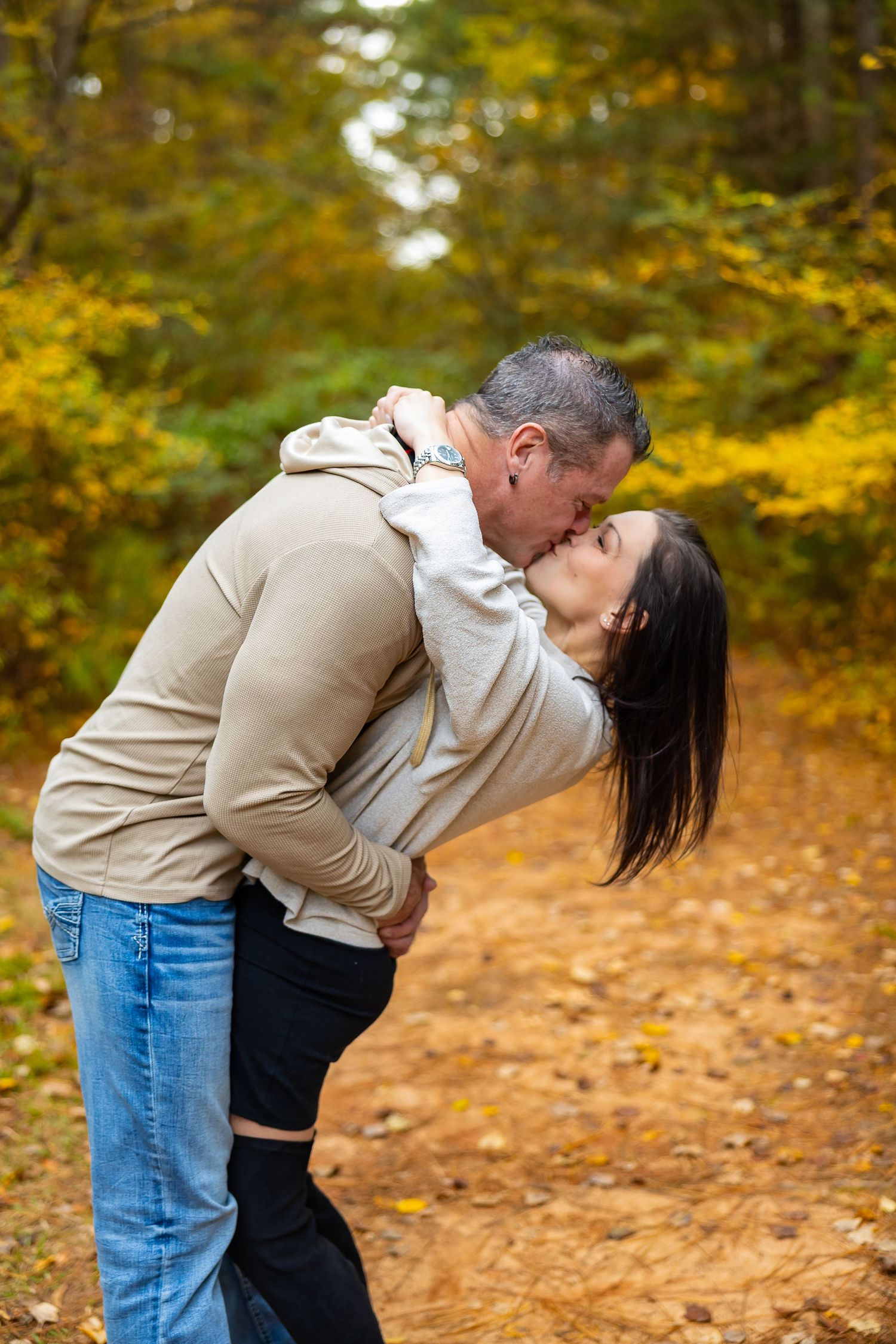 A romantic autumn scene shows a couple embracing and kissing on a path covered in fallen leaves with golden foliage above.