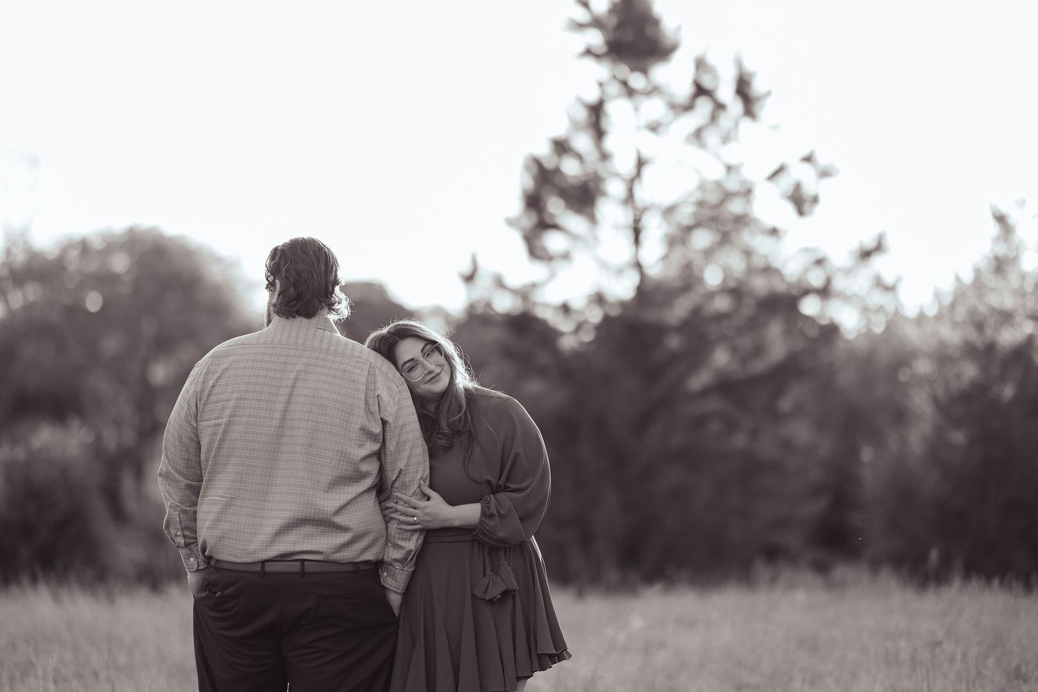 Black and white photo of couple embracing in field, woman resting head on mans shoulder.