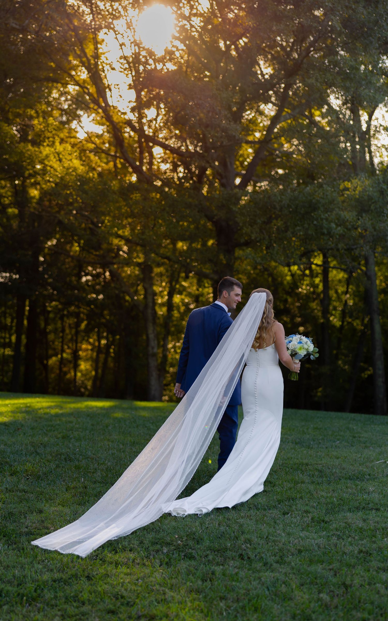 Bride and groom walking toward sunset through lush trees, her cathedral veil trailing gracefully on green grass.