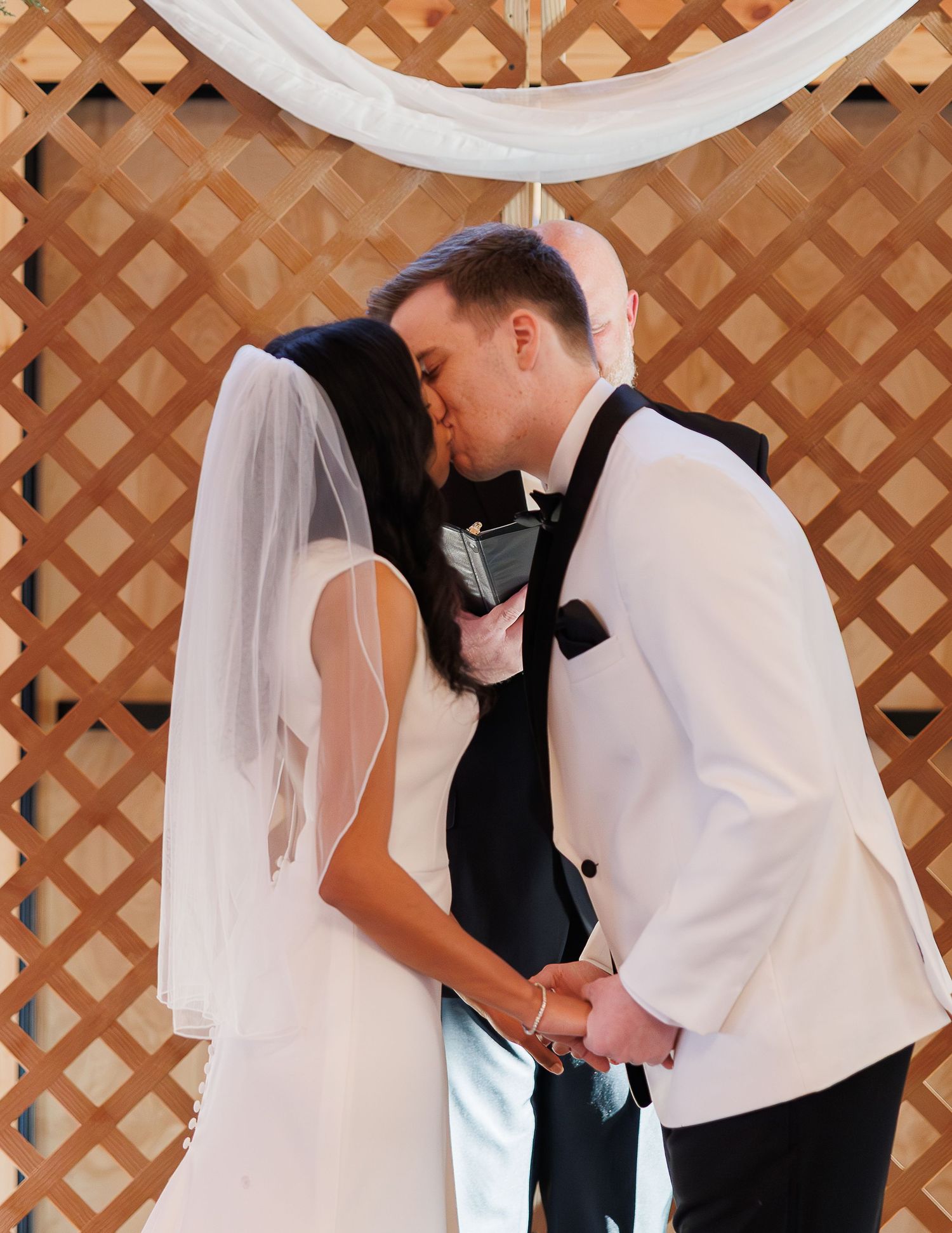Bride and groom share their first kiss at the altar during their wedding ceremony in front of a wooden lattice backdrop.