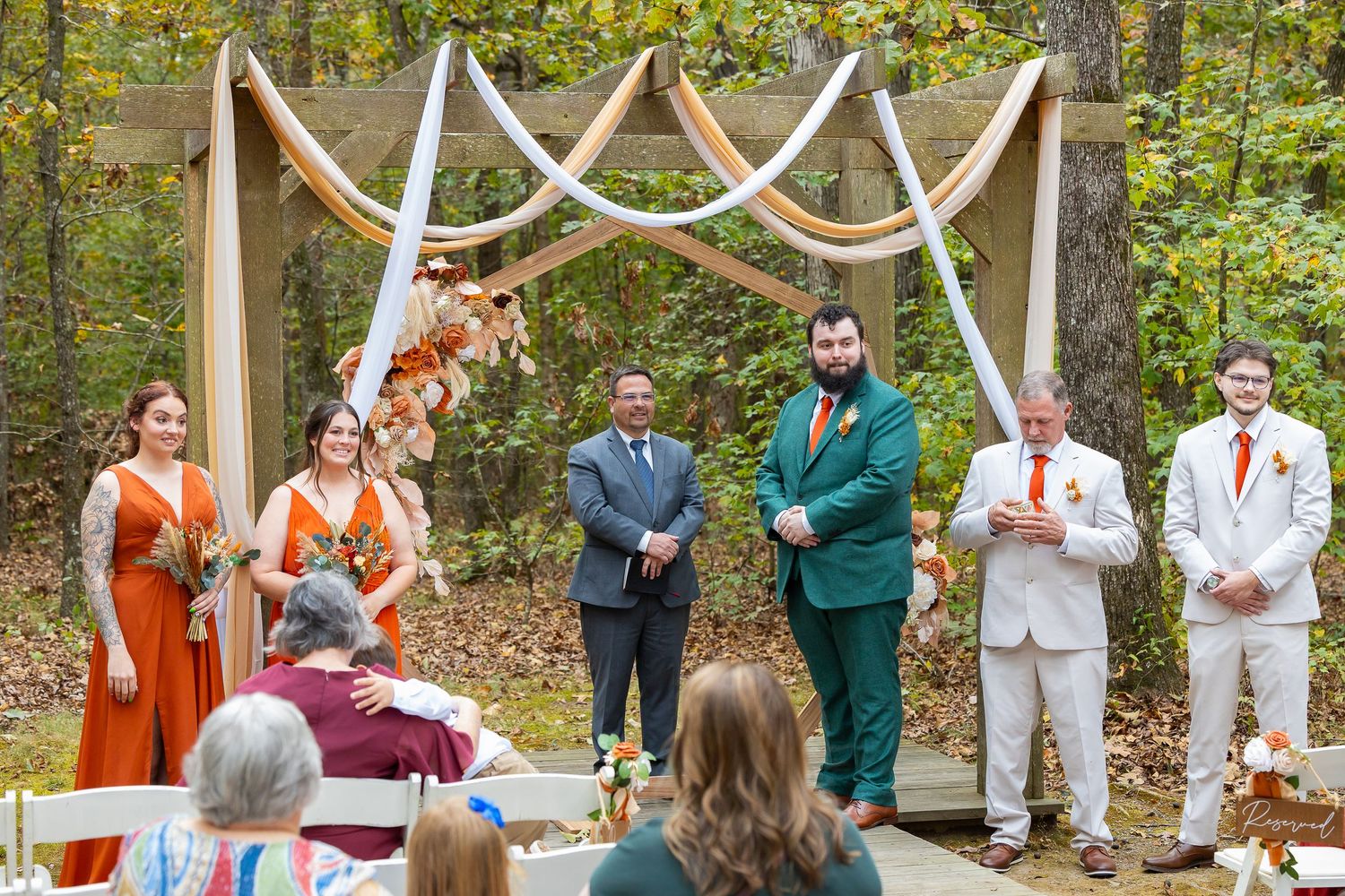 Outdoor fall wedding ceremony with wedding party in orange and green attire under a decorated arch in a wooded setting.