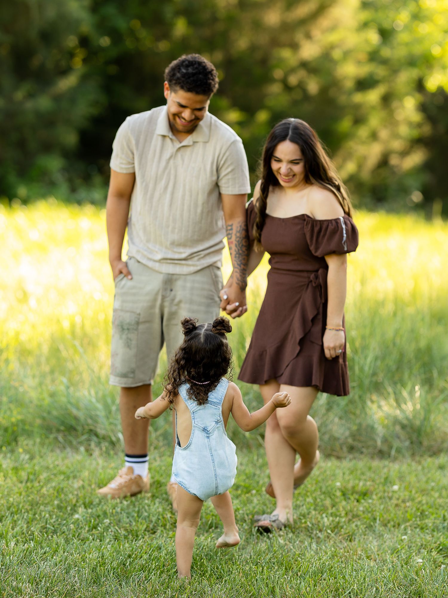 Family of three enjoying a sunny day in a green meadow, parents holding toddler girl's hands.