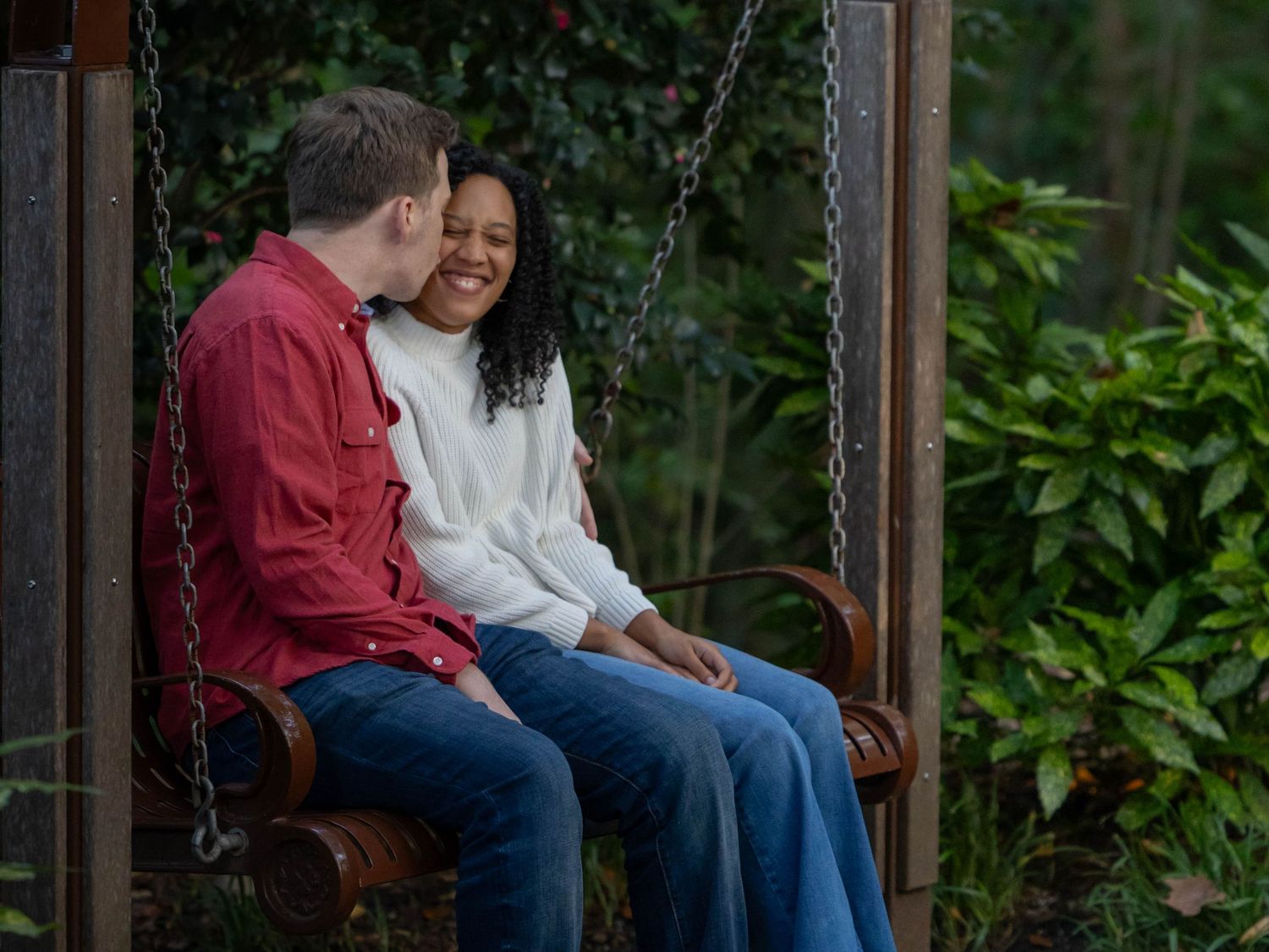 A couple sits close together on wooden steps surrounded by lush greenery in an outdoor setting.