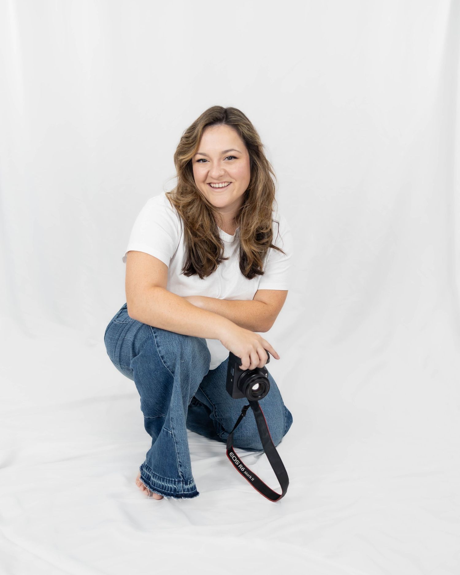 A person in blue jeans and a white top poses casually against a white studio backdrop.