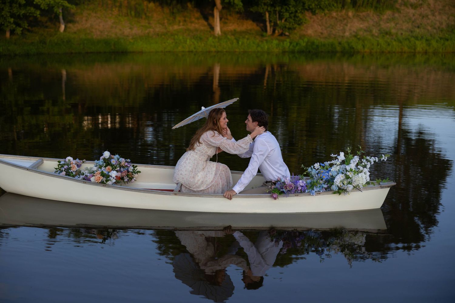 A romantic couple sits in a white rowboat on a tranquil lake surrounded by greenery and flowers.