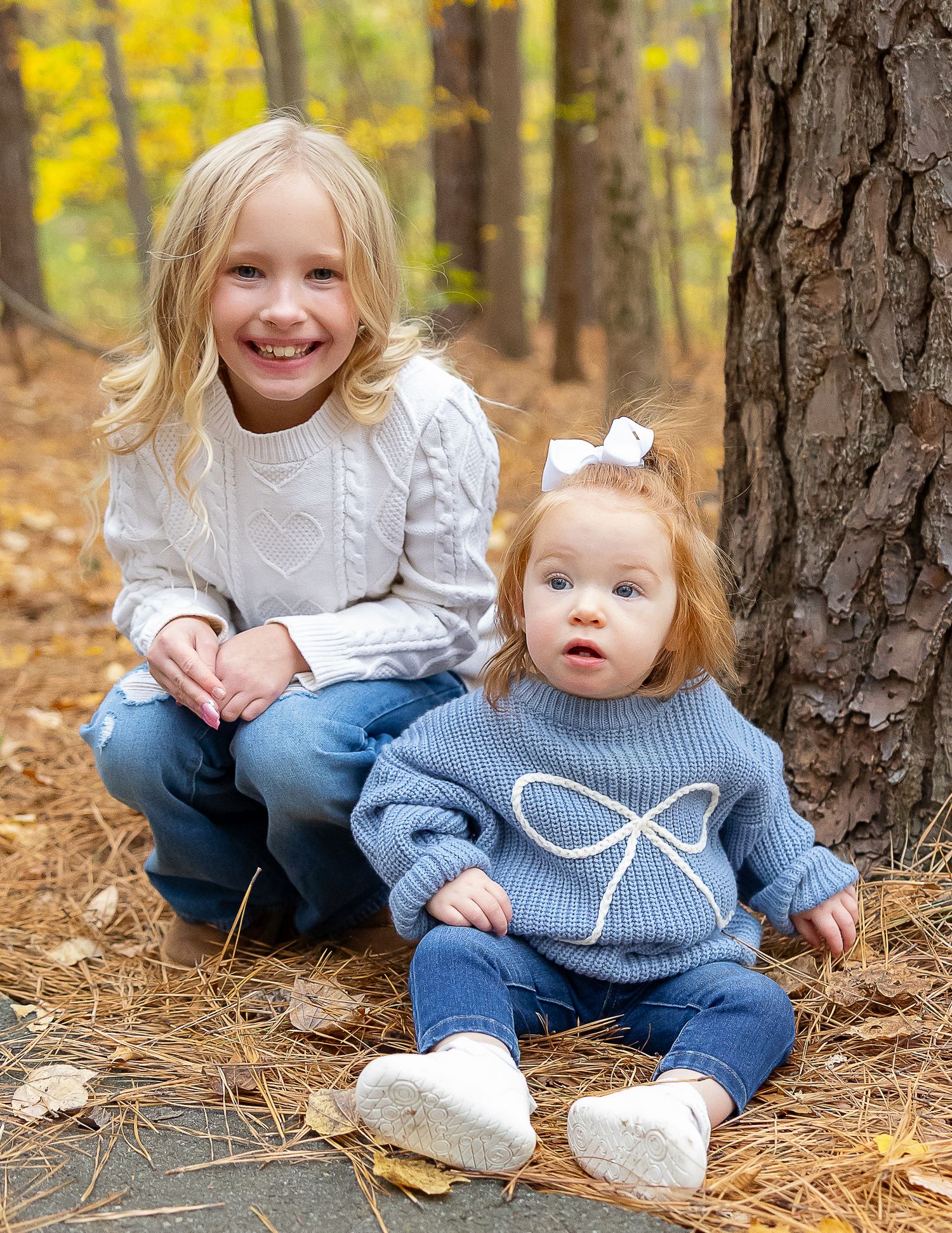 Two children sit together on fallen autumn leaves in a forest setting with golden yellow foliage in the background.