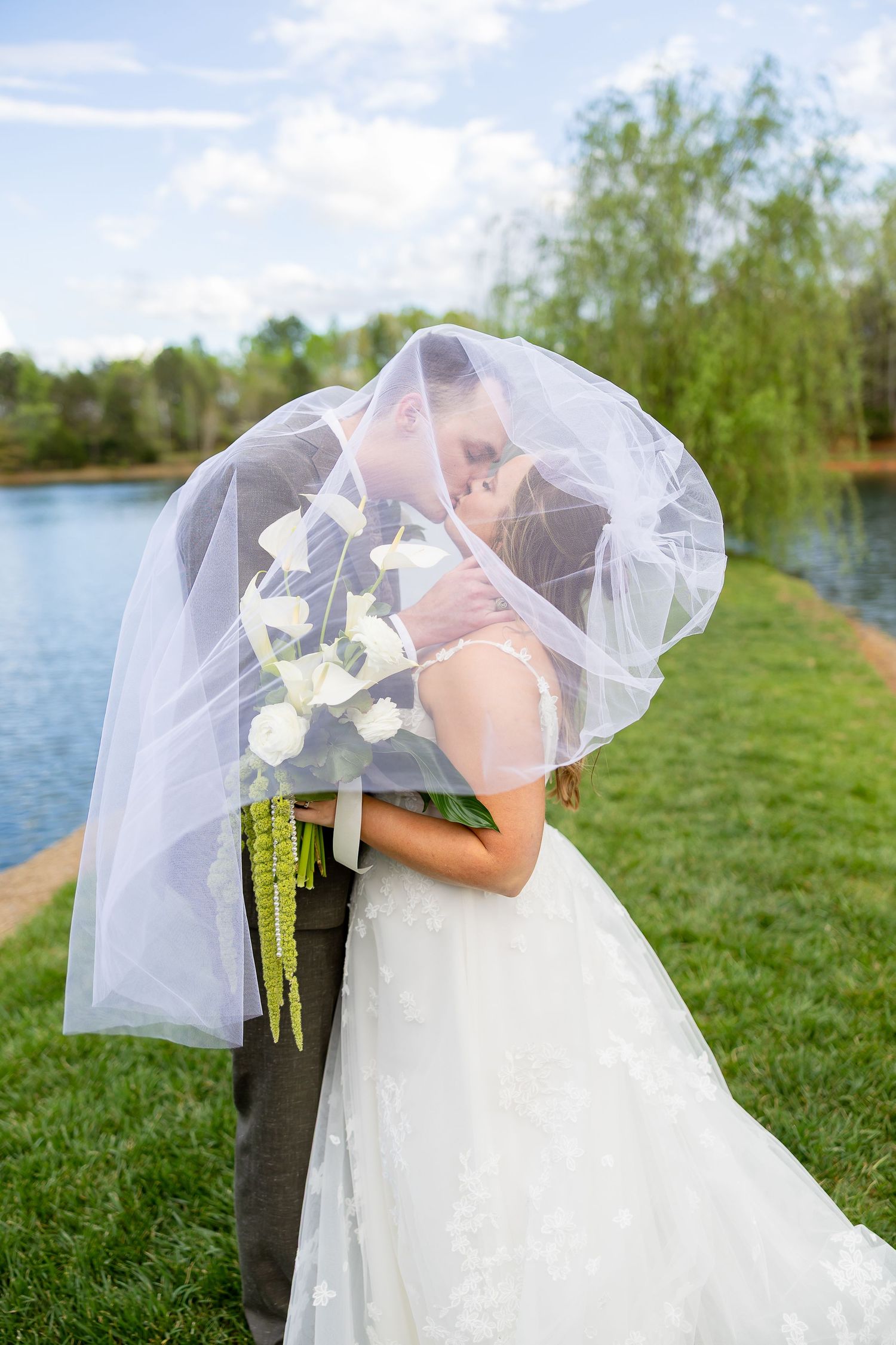 Bride and groom sharing a romantic kiss under a flowing white veil, holding white calla lily bouquet by a serene lake.