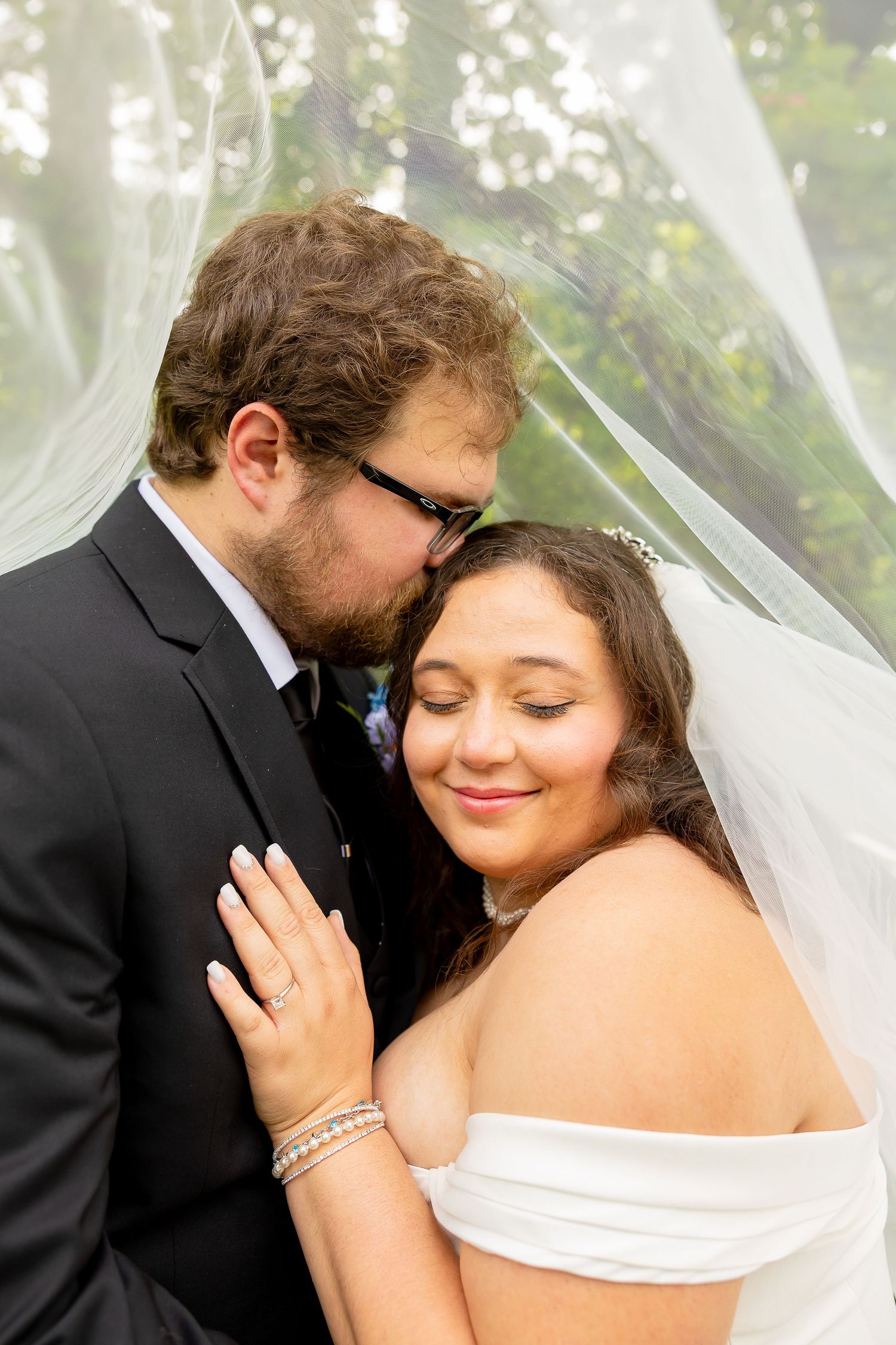 Groom kisses bride on the cheek under a sheer wedding veil as she smiles with eyes closed.