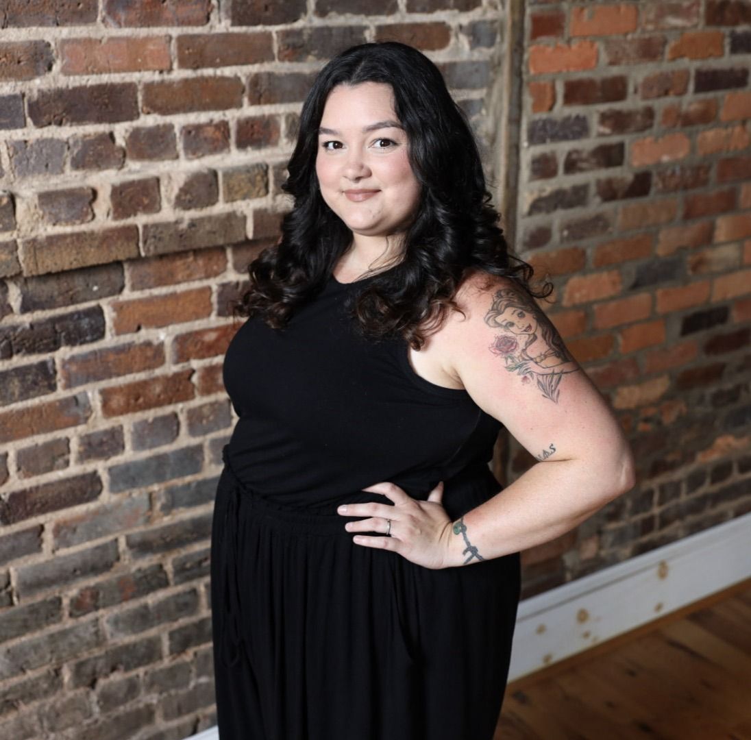 Woman in a black dress posing against a rustic brick wall backdrop.