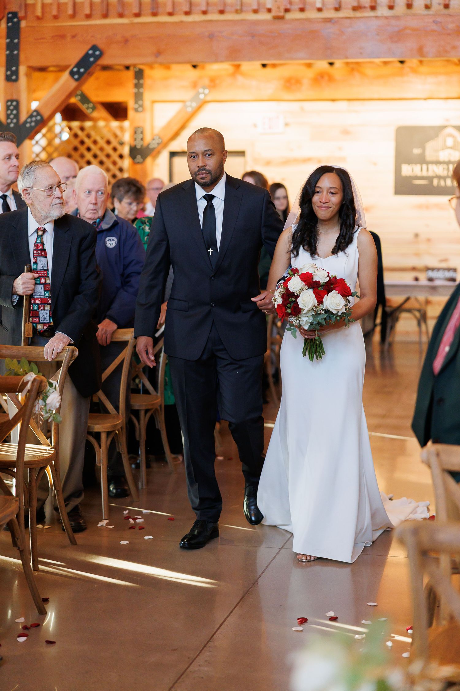 Bride walks down the aisle with her escort in a rustic barn venue, holding a bouquet of red and white roses.