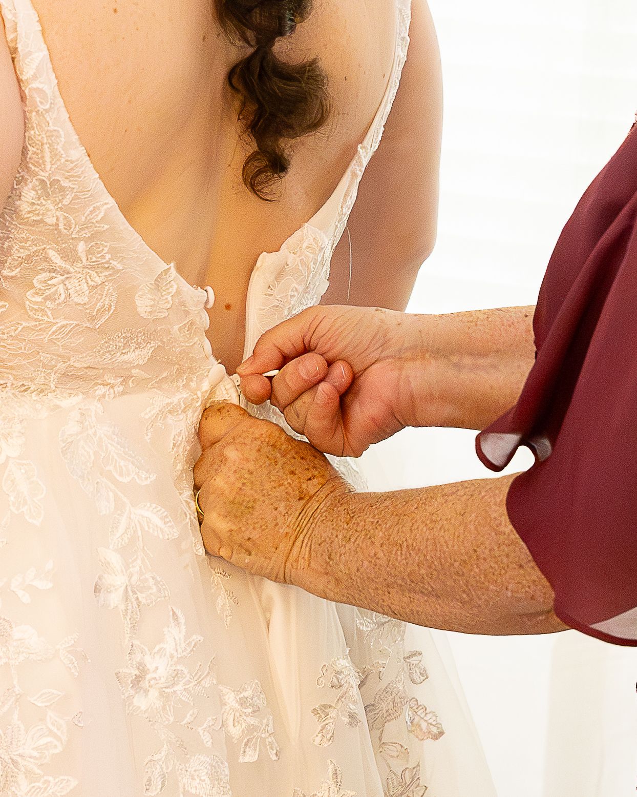 Elderly woman in burgundy dress fastening buttons on back of bride's white lace wedding gown.