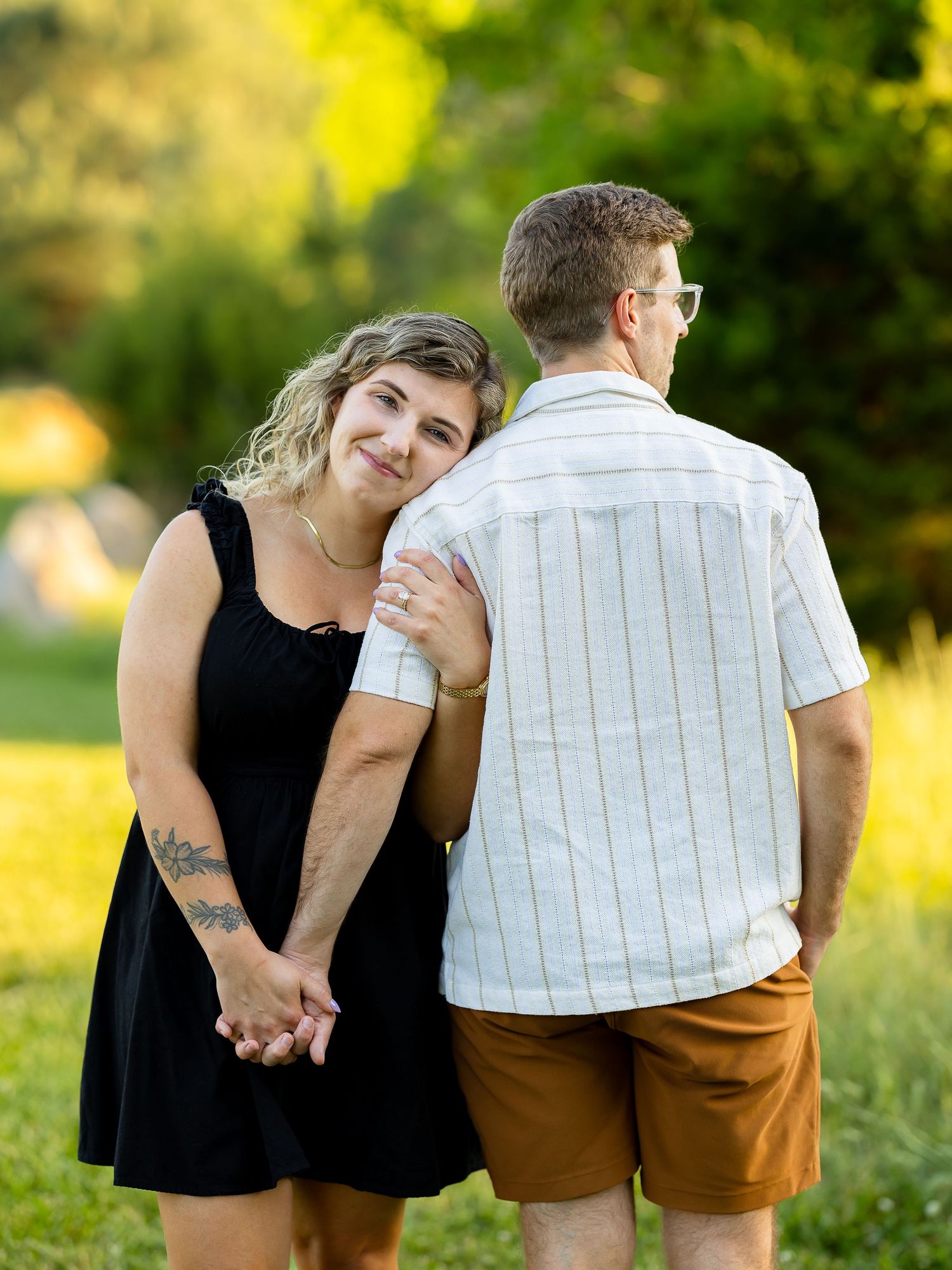 Couple holding hands outdoors, woman in black dress leaning on man's shoulder in golden sunlight.